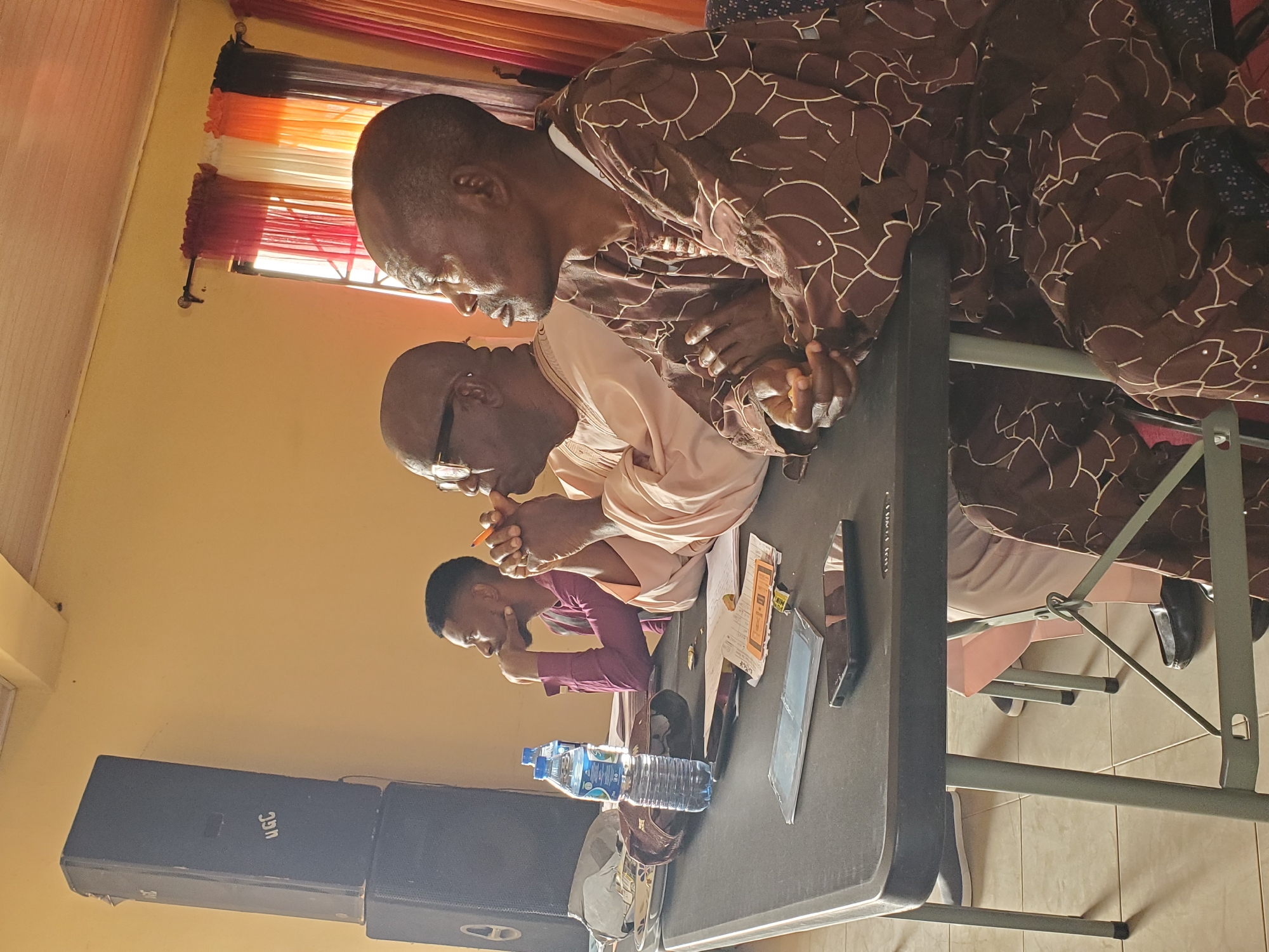 The image shows four men seated at a rectangular table, possibly indoors given the presence of a speaker system and curtains over a window. The man closest to the camera is wearing a brown patterned garment. Next to him is a man in a light peach robe wearing sunglasses. The third man is wearing a purple shirt, and the fourth man is partially obscured. A bottle of water, a cell phone, and documents are on the table. The men appear to be in conversation or discussion. The location is suggested to be Jos, Nigeria.