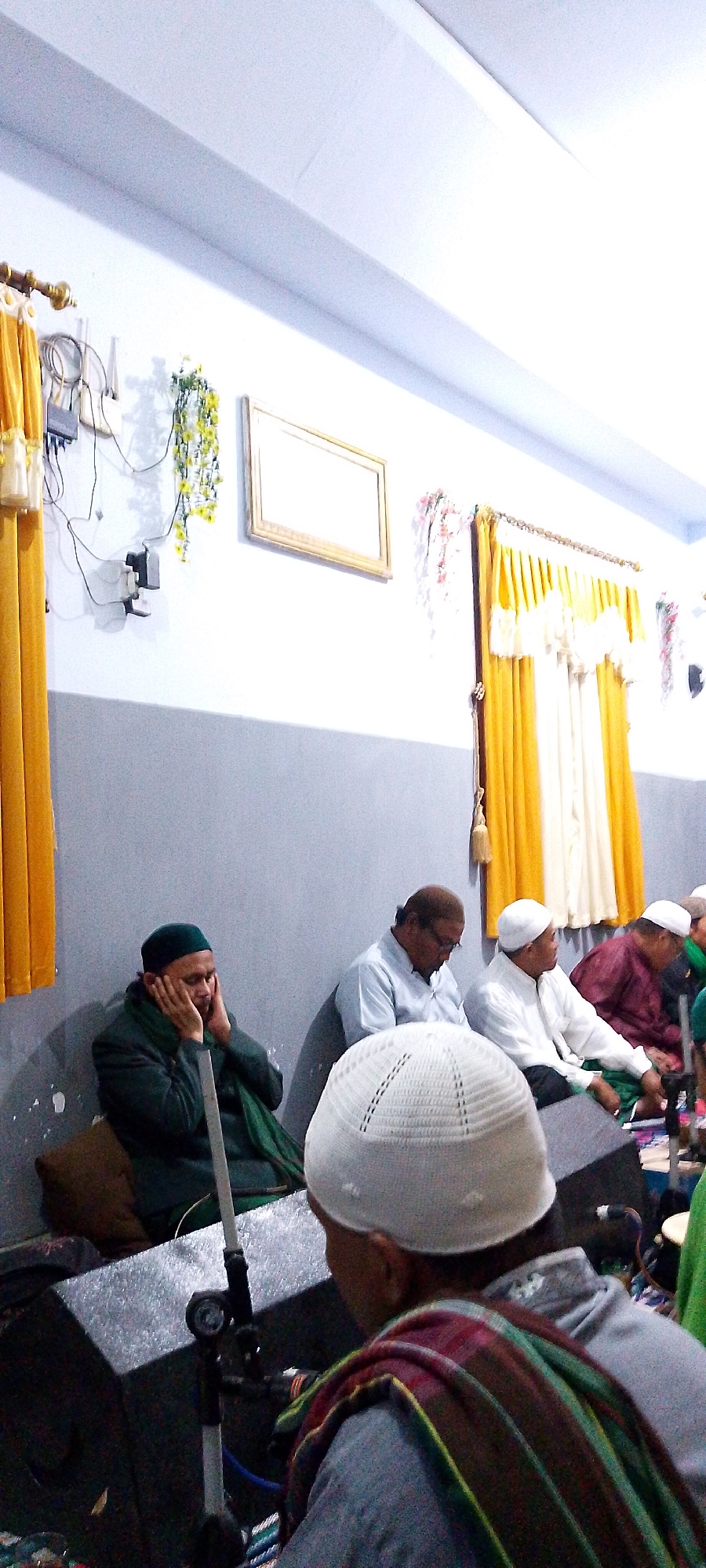 The image depicts an indoor gathering of several men, likely at a religious or community event, within the city of Oro-oro Ombo, Indonesia. The setting appears to be a room with white walls, a grey lower wall section, and a light-colored ceiling with beams. Yellow curtains with white trim are visible on the right side, and a decorative framed picture hangs on the wall.
In the foreground, a man wearing a white kufi and a colorful striped scarf over a grey garment is seen from the back, facing away from the camera. To his left, a man in a dark green robe and a matching kufi has his hands on his face, appearing thoughtful or perhaps sleeping. Next to him, another man wearing a light blue long-sleeved shirt and glasses is also seated. Further to the right, a man in a white kufi and shirt is visible, and beyond him, a man in a maroon robe is seated. The men are seated on the floor or on low platforms, with a large black speaker system in the immediate foreground. The overall atmosphere suggests a moment of reflection or prayer within a communal setting.