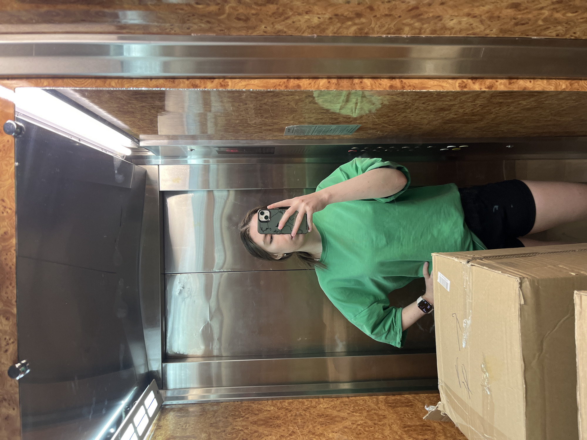 A young woman is taking a selfie in what appears to be an elevator. She is wearing a green t-shirt and black shorts. In her left hand, she holds a phone up in front of her face. Her face is partially obscured by the phone. On her left wrist she has a wristwatch. The elevator's interior consists of polished metal walls and wood-paneled ceiling and floor. Several large cardboard boxes are in the elevator with her. Based on the style and detailing of the elevator, as well as the wood paneling, it may be in Moscow, Russia.