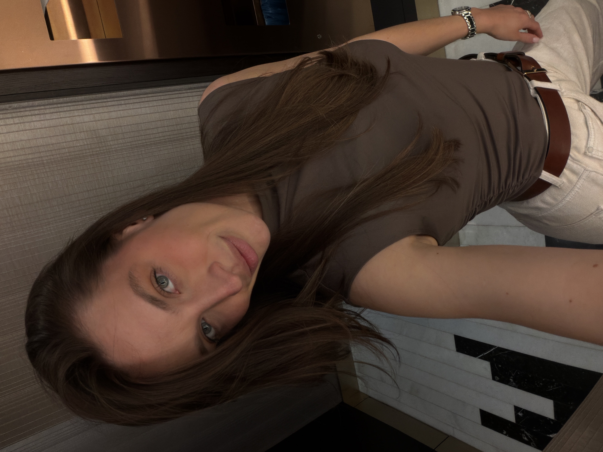 A medium-shot angled selfie of a woman with fair skin and long brown hair. She is wearing a brown t-shirt, beige pants with a brown belt, and a silver wristwatch. The background consists of a tiled wall and floor with a mix of light and dark tiles. The location is potentially a bathroom or similar space with contemporary design. It's not possible to determine the location in Budva, Montenegro without additional information.