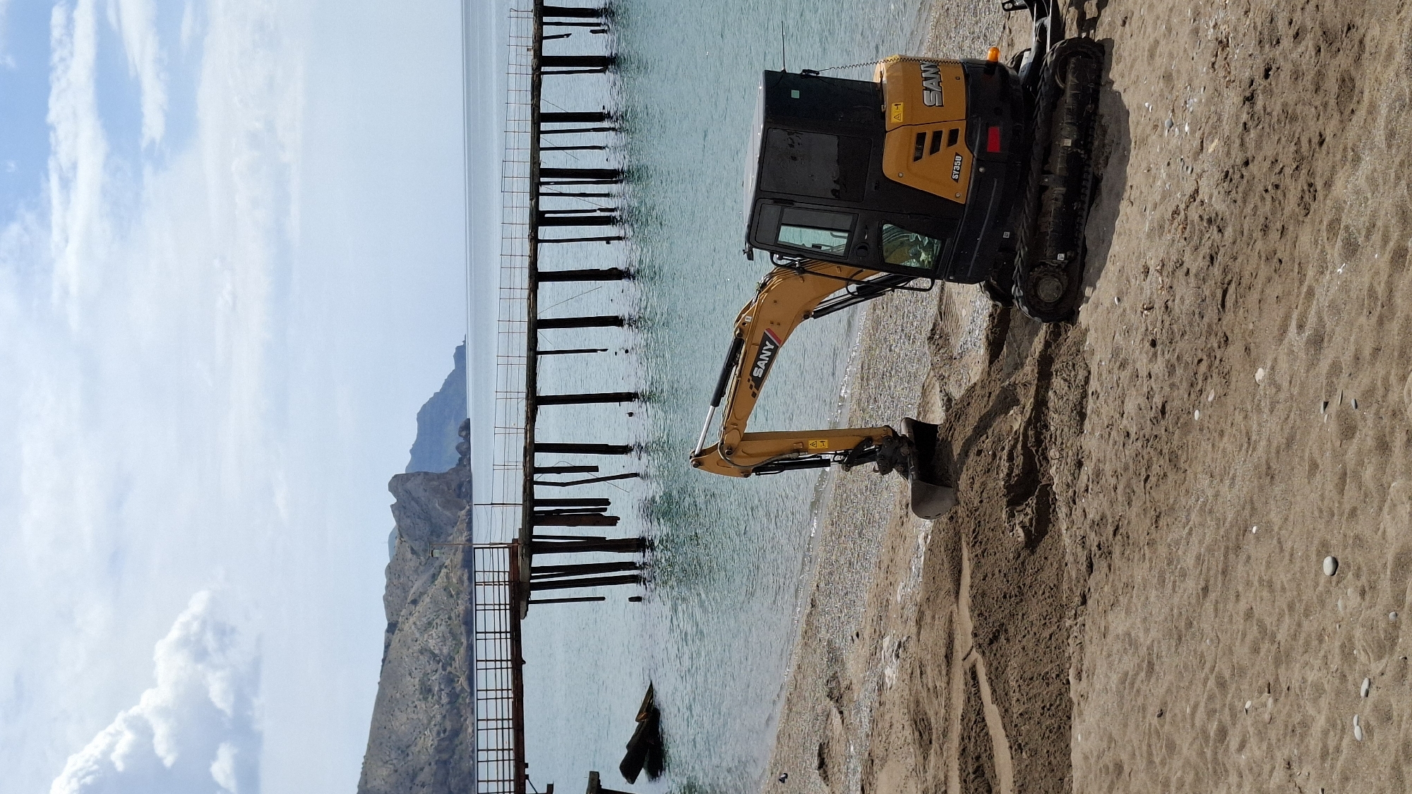 The image shows a SANY SY35U excavator on a sandy beach. The excavator is positioned near a partially submerged pier or jetty constructed of wooden pilings and metal framework. In the background, a mountain range is visible. The sky is light blue with scattered clouds. The location is Unknown, Unknown.