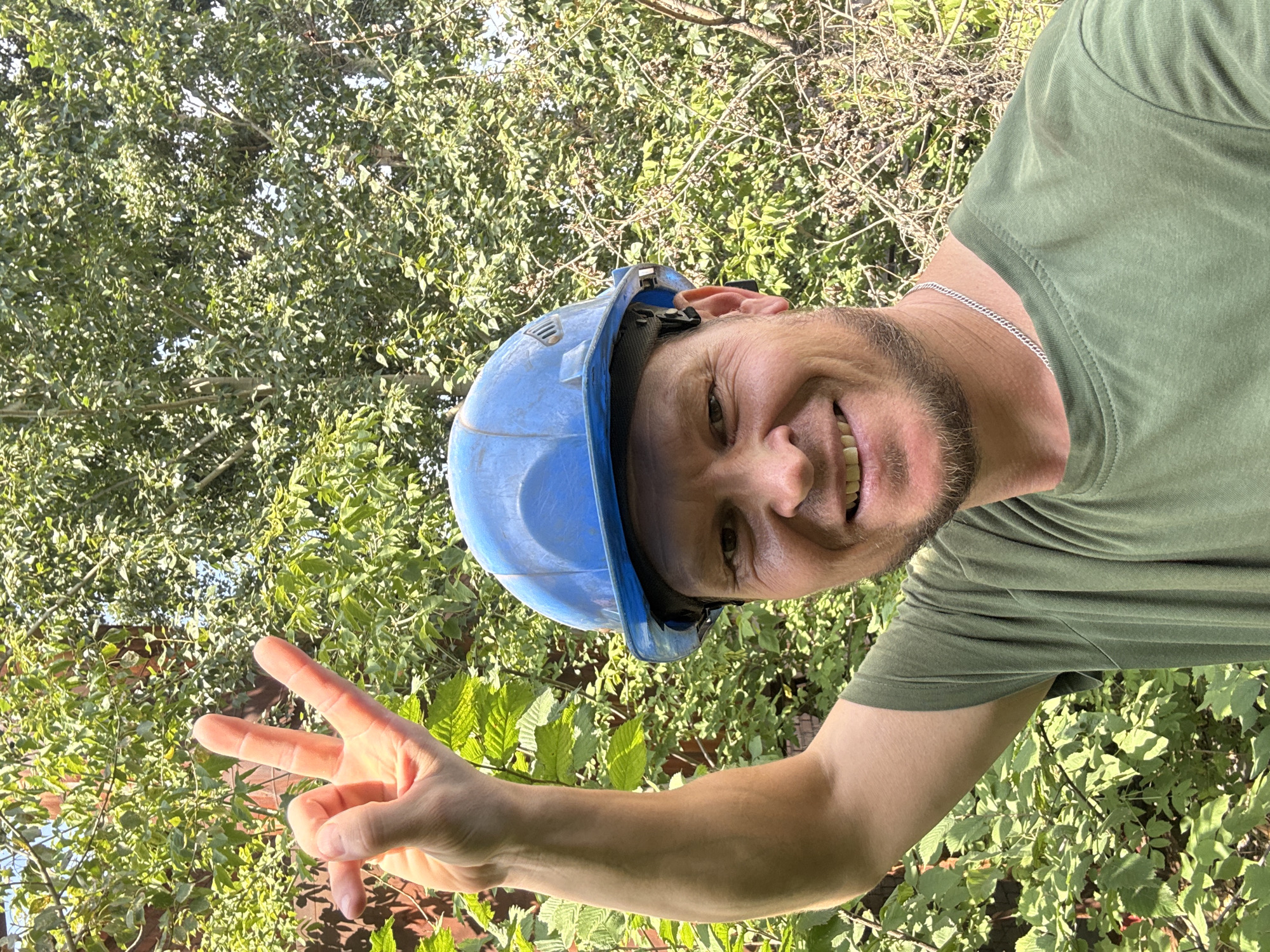 A man wearing a blue hard hat and a green t-shirt is outdoors, with lush green foliage in the background. His right arm is raised, and he is holding up three fingers in a peace sign. He has stubble on his chin and is smiling. The scene appears to be a sunny day, with shadows cast by the trees. The location is described as the city of Oskemen, Kazakhstan.