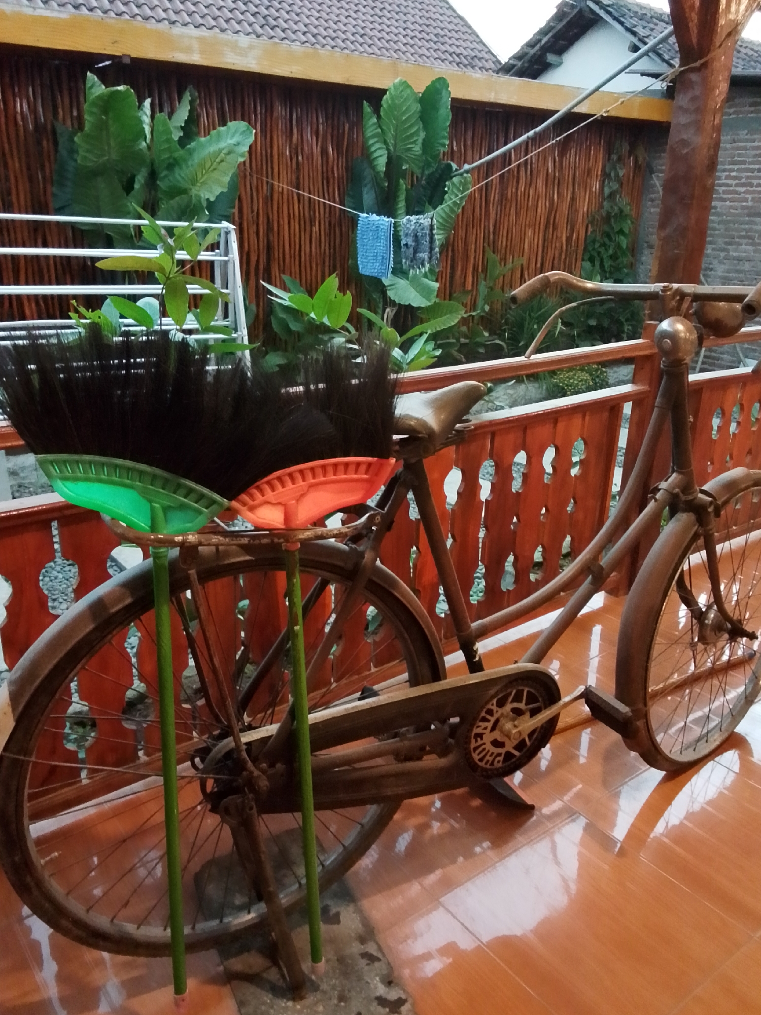This image depicts a rustic scene featuring an old bicycle with two brooms attached to its frame. The bicycle is leaning against a wooden railing with decorative cutouts. Behind the bicycle, a wall constructed of vertical wooden poles is visible, with lush green plants growing in front of it. A clothesline with two small towels is strung across the scene. The ground is covered in glossy orange tiles, reflecting the light. The overall setting suggests an outdoor area, likely a backyard or patio, in Mojotengah, Indonesia. The bicycle appears to be made of dark metal and shows signs of age. The brooms have dark bristles and brightly colored plastic heads, one green and one orange, held by green poles. The background includes a hint of roofing tiles and brickwork, suggesting the presence of buildings.