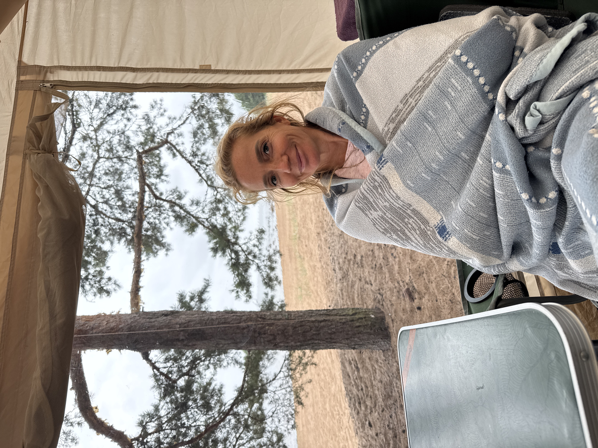 A Caucasian woman with blonde hair is wrapped in a grey and white striped blanket, seated in a tent. She is wearing a light pink shirt and a necklace. The tent is open, revealing a sandy beach and a pine tree in the background under a cloudy sky. A green table with a metallic edge is visible to the left of the woman.