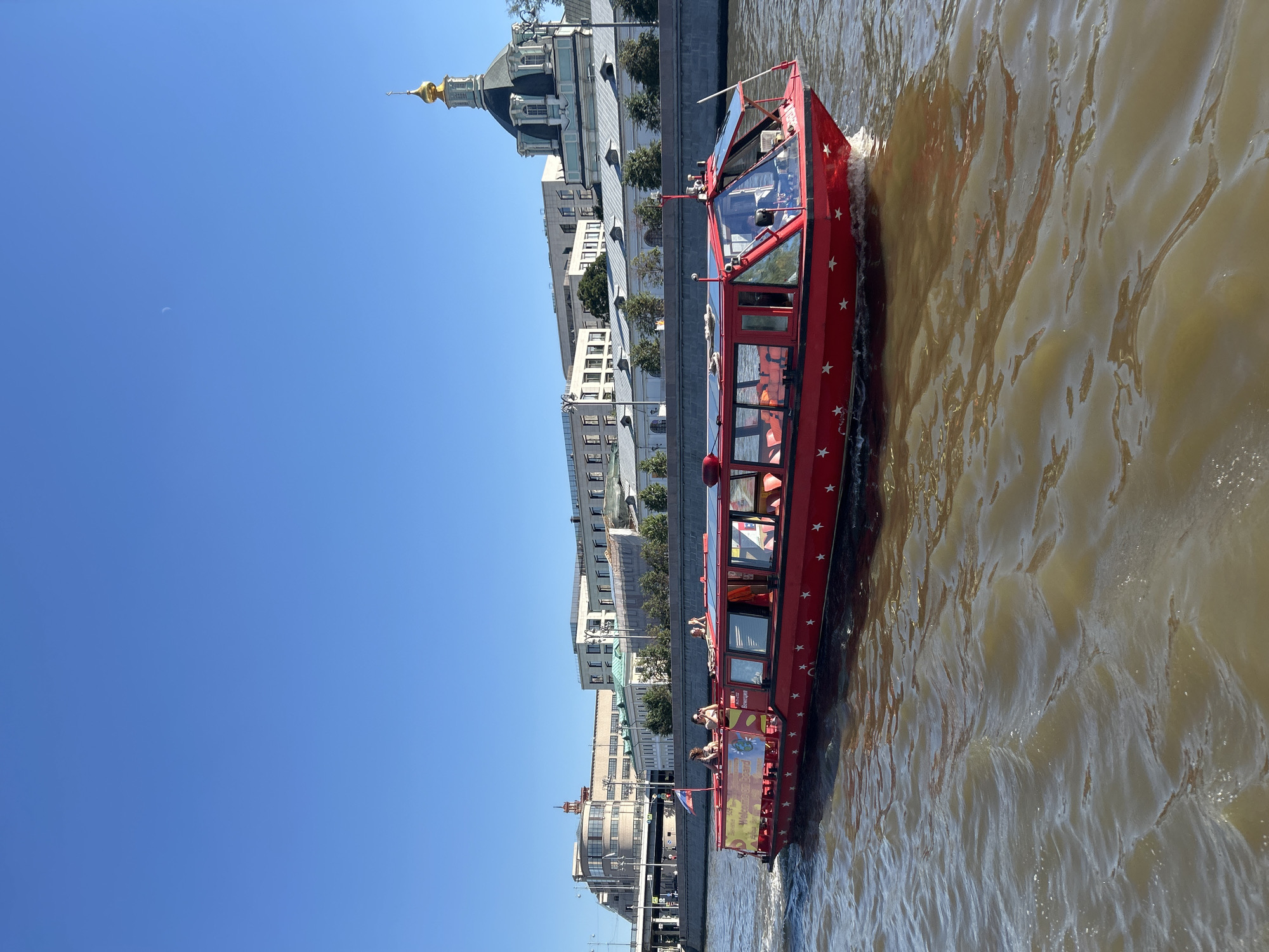 This image captures a bright, sunny day in Moscow, Russia, likely on the Moskva River. A vibrant red tour boat, adorned with white stars and the text "Welcome On Board," cruises through the water, creating ripples. Several passengers are visible on the deck of the boat, enjoying the excursion. In the background, a line of historic buildings and a domed church with a golden cupola line the riverbank. The sky is a clear, cloudless blue, indicating good weather conditions.