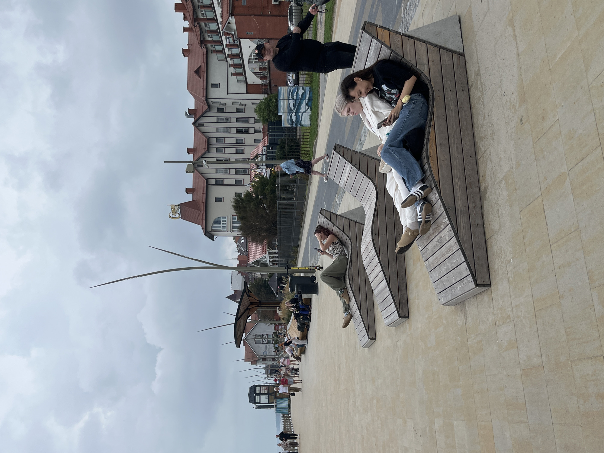 The image depicts a seaside promenade in Zelenogradsk, Russia, under a cloudy sky. Several people are visible in the scene. In the foreground, two women are relaxing on modern wooden chaise lounges. One woman is looking at her phone, while the other leans her head back. Further back, another woman is lying on a chaise lounge, also looking at her phone. In the background, several buildings line the promenade, featuring red-tiled roofs and white facades. People are walking along the promenade, and there are decorative streetlights and benches. A man in a black shirt and cap stands near the edge of the promenade, appearing to observe the surroundings.
