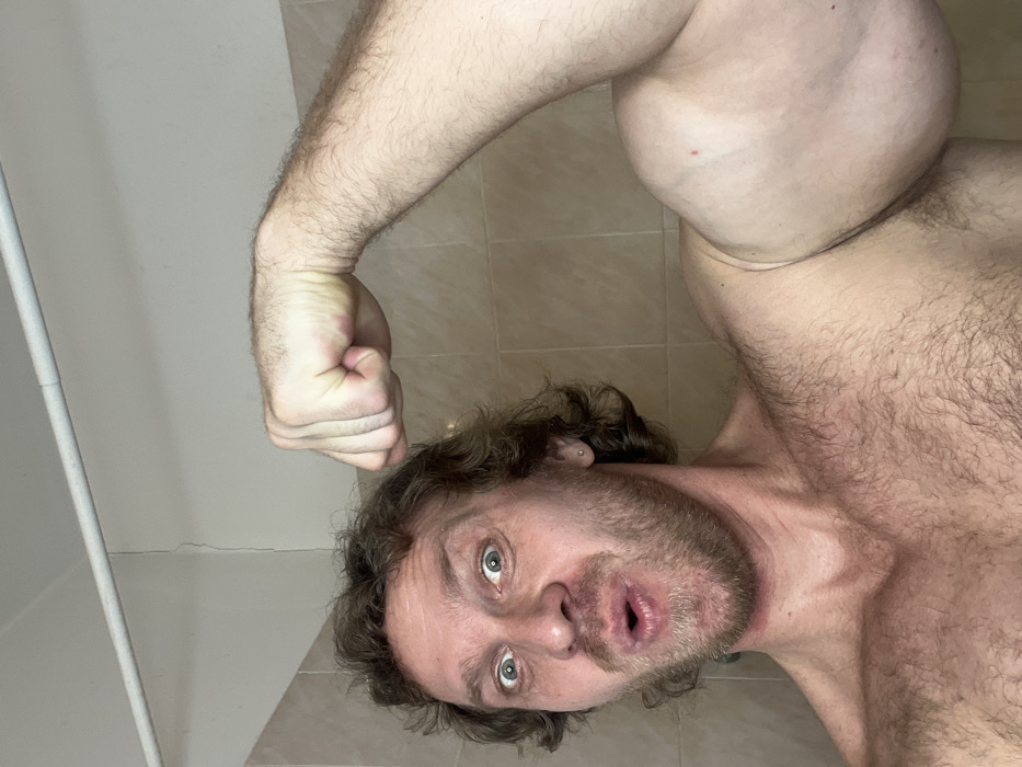 The image displays an adult male, shirtless, posing indoors, reportedly in Saint Petersburg, Russia. The man has curly brown hair, a light brown beard, and blue eyes that are wide open and directed towards the viewer. His mouth is open in a circular shape, and he has a silver stud piercing in his left earlobe. He is flexing his right arm, with his hand clenched into a fist, showcasing a significantly developed and rounded right bicep. His chest and shoulders are visible and covered in body hair. The background consists of light beige square wall tiles extending from below the man's shoulders, above which is a plain white wall. A distinct vertical crack is visible on the white wall. A white pipe runs horizontally across the top of the white wall.