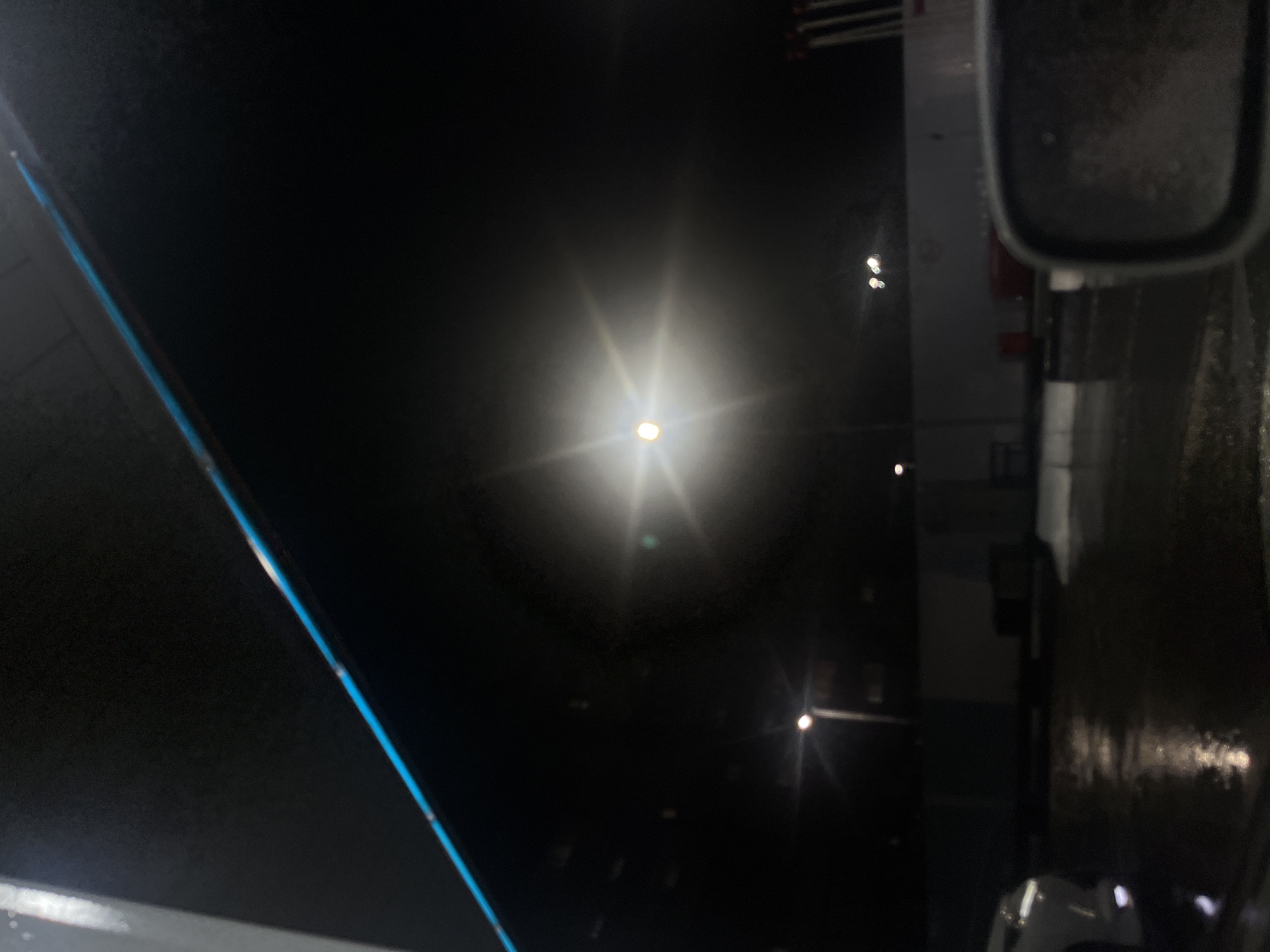 The image depicts a dark, nighttime scene, possibly at a parking lot or an outdoor area. The dominant feature is a bright, diffused light source in the center, creating a halo effect. A blue-tinged line is visible along the top edge, suggesting an artificial structure or lighting. In the lower left, the rear of a white vehicle is partially visible, reflecting on a wet surface. The ground appears wet, with reflections of lights indicating recent rain or moisture. In the background, there are indistinct structures and what appear to be multiple light poles, some with multiple lights. The overall atmosphere is gloomy and wet, characteristic of a city environment at night, potentially in Belogorsk, Russia, given the context. There are no people or specific identifiable landmarks visible.