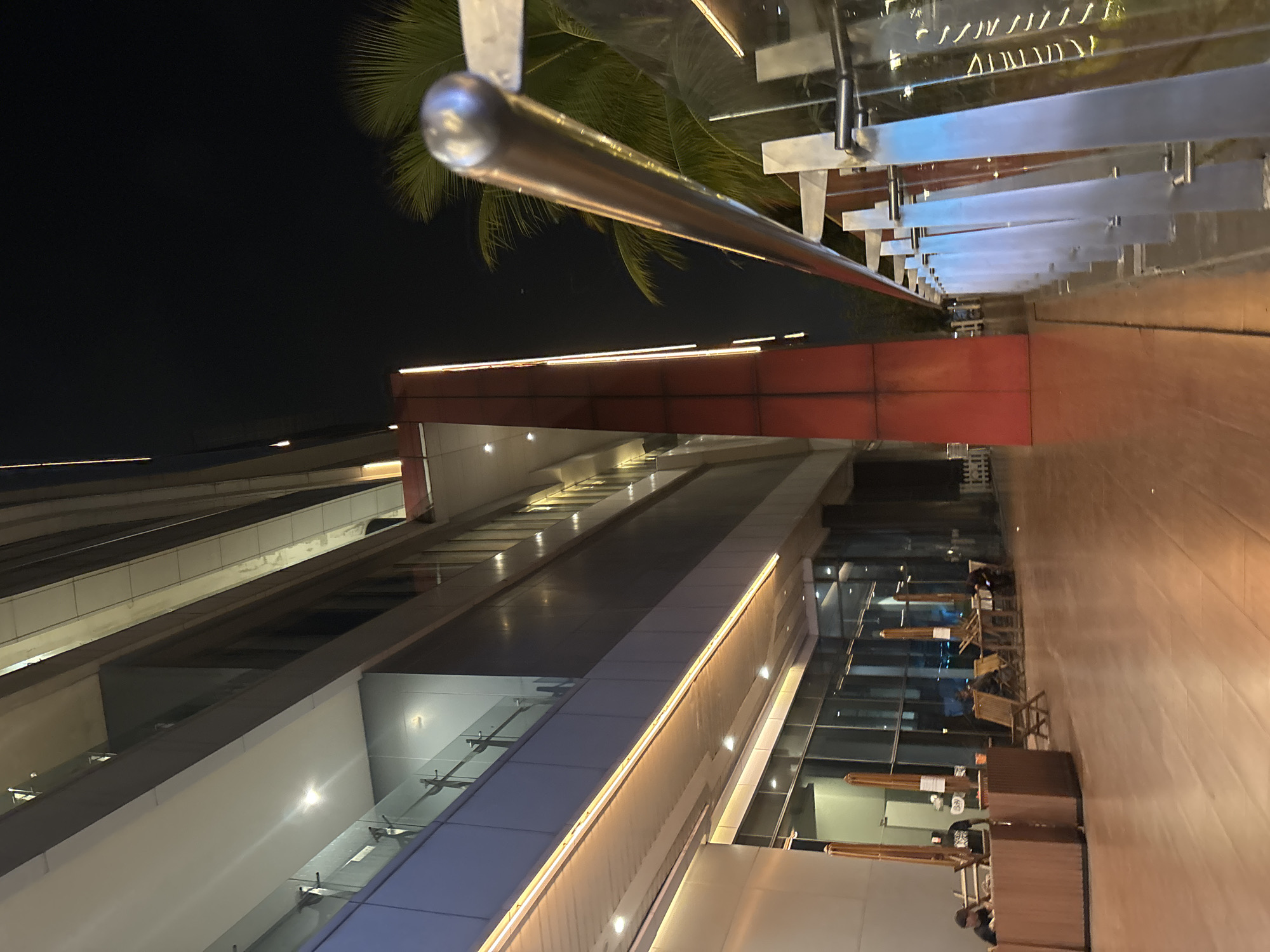 The image captures an outdoor scene at night in North Jakarta, Indonesia, focusing on the exterior of a modern building with a predominantly glass facade and wood accents. The building features multiple levels with balconies and illuminated walkways. A prominent red, rectangular architectural element extends vertically on the building's exterior. In the foreground, a polished metal railing with glass panels lines a walkway, reflecting the building's lights and the night sky. Palm fronds are visible in the background.
On the lower level of the building, large windows reveal an interior space that appears to be a seating area. Several wooden chairs and tables are arranged on a wooden floor, with some empty and others occupied by individuals. Two people are seated at a wooden counter or desk on the left side of the frame, possibly at a reception or service area. Umbrellas are positioned near the seating area. The overall impression is that of a public space, possibly a commercial establishment or a hotel lobby, at dusk or nighttime.
