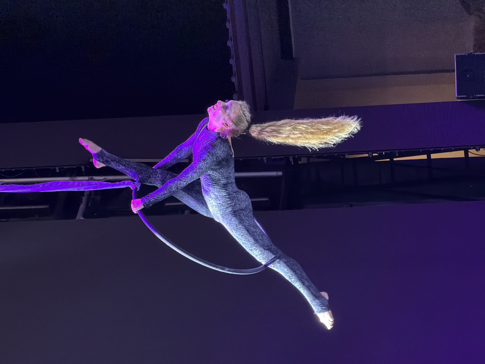 An aerial performer, with blonde hair pulled into a ponytail and wearing a patterned grey costume, is suspended mid-air by a purple rope and a hoop. The performer is upside down, with their legs extended and crossed. Their body is positioned in a graceful, athletic pose. The background consists of a dark, textured surface and purple lighting, suggesting a stage or performance venue. The location is specified as Manavgat, Turkey, though no specific landmarks are visible in the image to confirm this.
