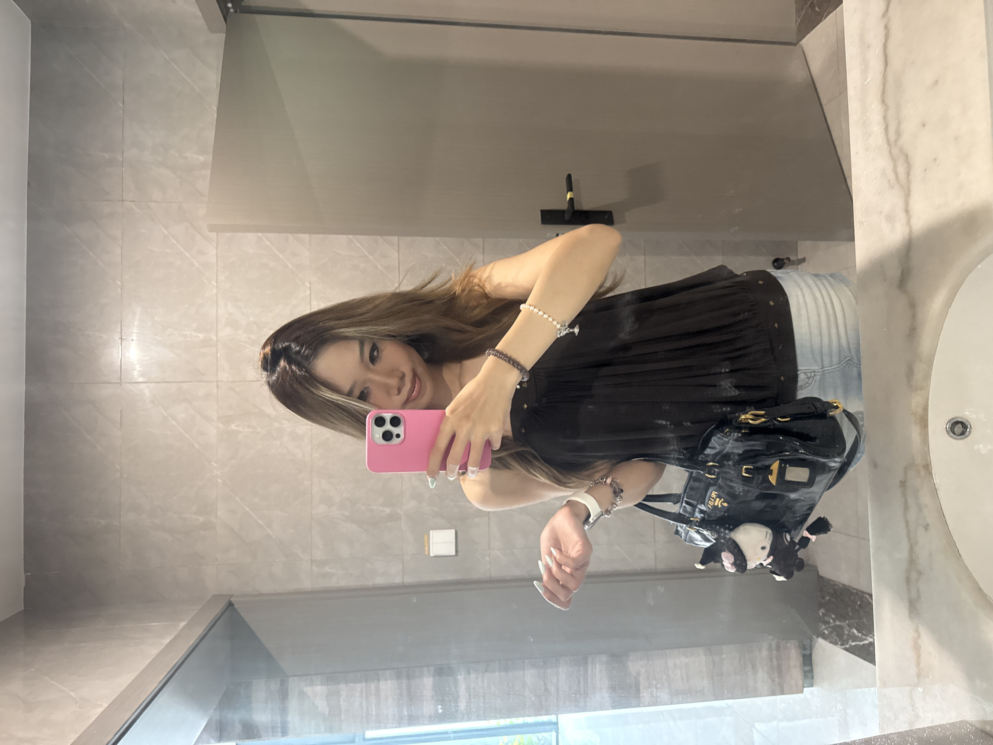 A young woman is taking a selfie in a bathroom. She is holding a pink smartphone in her left hand and looking at the screen. She has long brown hair and is wearing a black top and light blue denim shorts. She also has bracelets on her wrist. Beside her is a black purse with a black and white keychain attached. The background shows a modern bathroom with light gray tile walls, a glass shower enclosure, and a white marble countertop with a sink. The reflection in the mirror suggests the presence of a window. While I am unable to identify the precise location, the setting is within a modern bathroom environment in an urban area, possibly the Nan'an District, China.