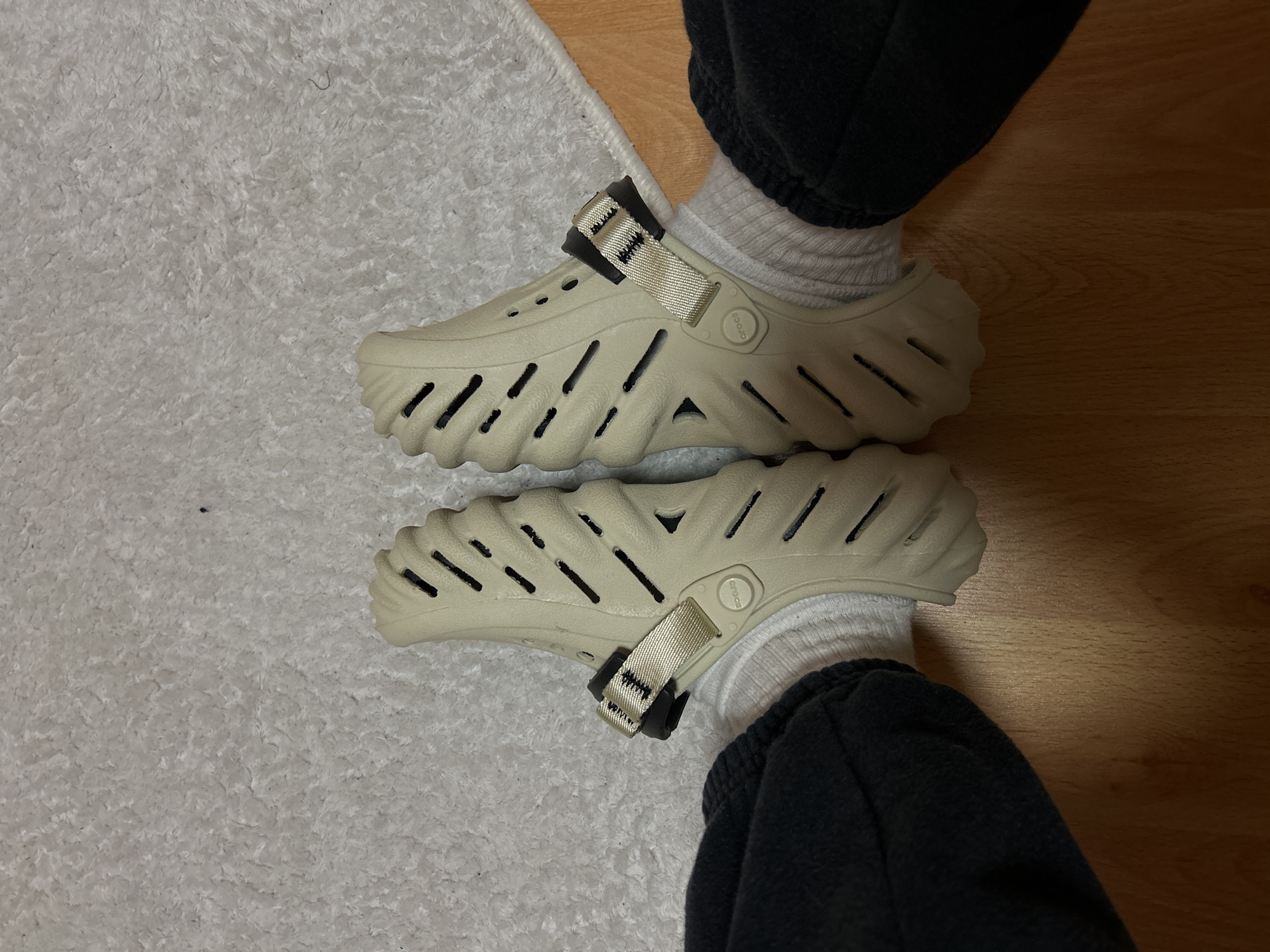The image shows a pair of feet wearing beige Crocs Echo clogs with white socks. The feet are positioned on a surface that is partially covered by a white shag rug and partially by a wooden floor. The person is also wearing dark sweatpants. The photograph does not contain information about the city of Goce Delcev, Bulgaria.