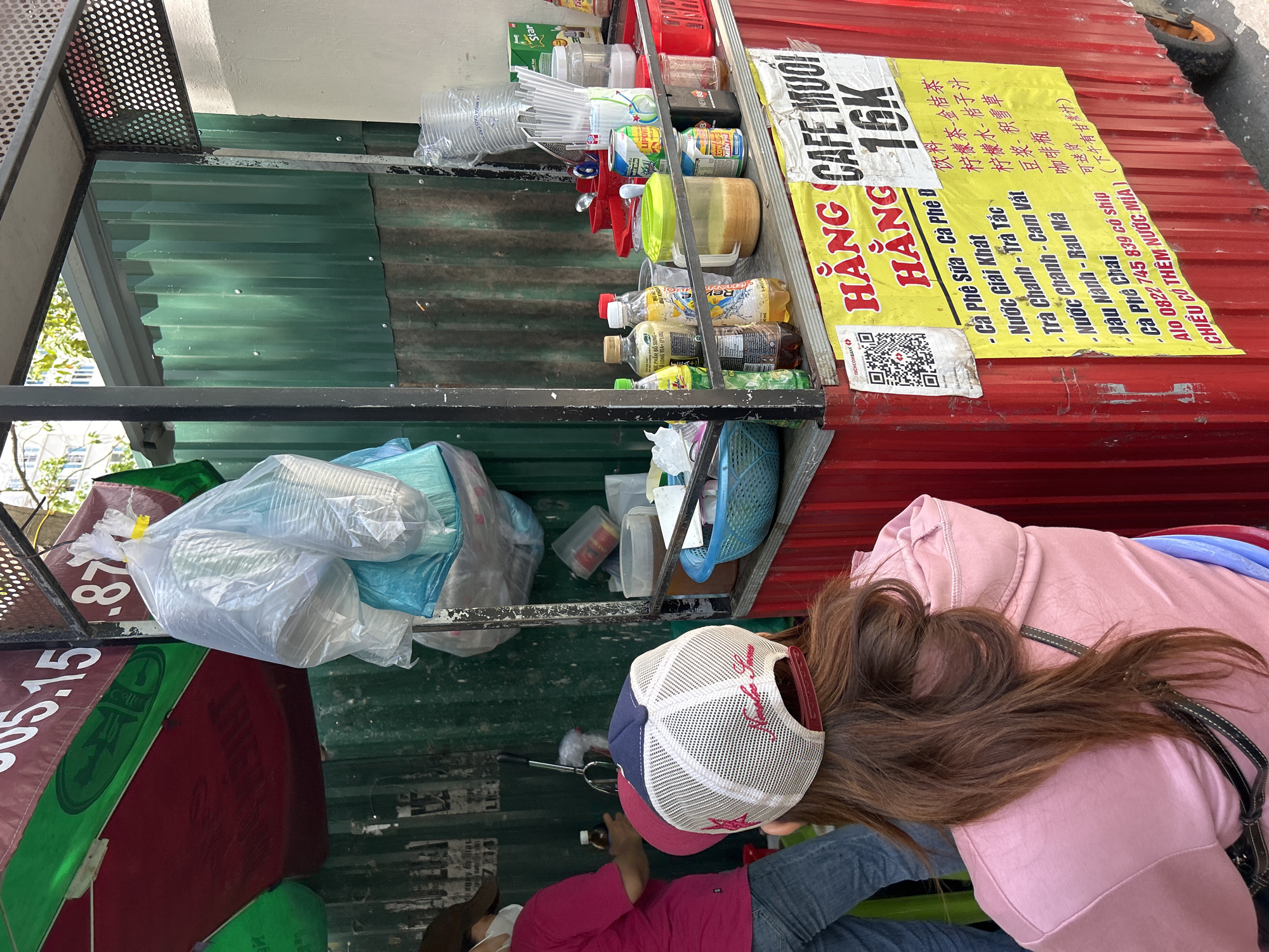 A street food vendor, likely in Bac Nha Trang Ward, Vietnam, is depicted from a slightly elevated perspective. The vendor's cart is constructed of corrugated metal painted red and green, and contains a variety of items, including packaged drinks, containers, and disposable cups. A yellow sign displays the name "HÃNG CAFE MUỐI HẰNG 16K" and lists beverage offerings in Vietnamese, with a QR code. A person is bending over the cart, possibly making a purchase or interacting with the vendor. The person is wearing a pink hoodie, jeans, and a baseball cap.