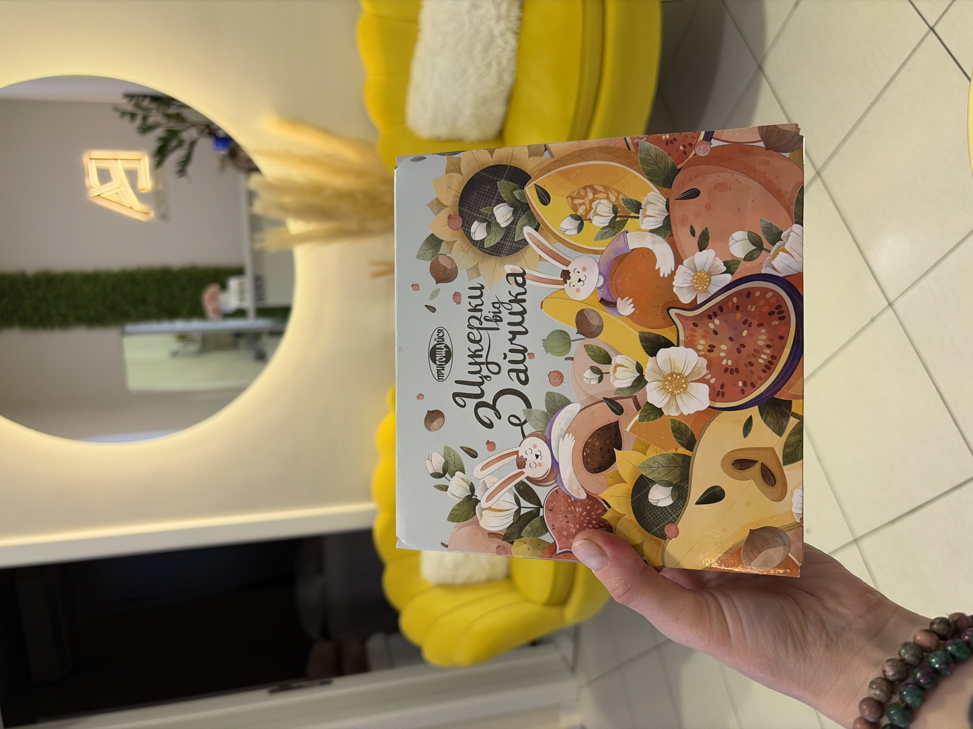 A hand holds a box of Ukrainian candy in front of a yellow chair in an indoor location, possibly in Budva, Montenegro. The box features an illustration of fruits and a cartoon rabbit. In the background, a mirror reflects an illuminated sign and greenery, creating a modern, decorated interior. The floor is tiled, and a shelf is visible below the mirror. The person holding the box wears beaded bracelets on their wrist.