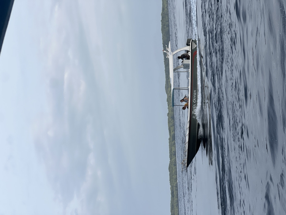 A dark-hulled motorboat, identified by the name "Twin Ocean" on its side, traverses the dark blue, rippled waters near Toyapakeh, Indonesia. The vessel, equipped with a white overhead canopy, is actively generating a white wake and bow spray as it moves. Three individuals are present on board: one operator, dressed in dark clothing, is positioned at the helm on the boat's starboard side. Two additional individuals, presumably passengers, are visible seated or standing mid-ship on the port side. The background features a distant, low-lying landmass covered in dense green foliage, consistent with a coastline or island, under a uniformly overcast, light grey sky.