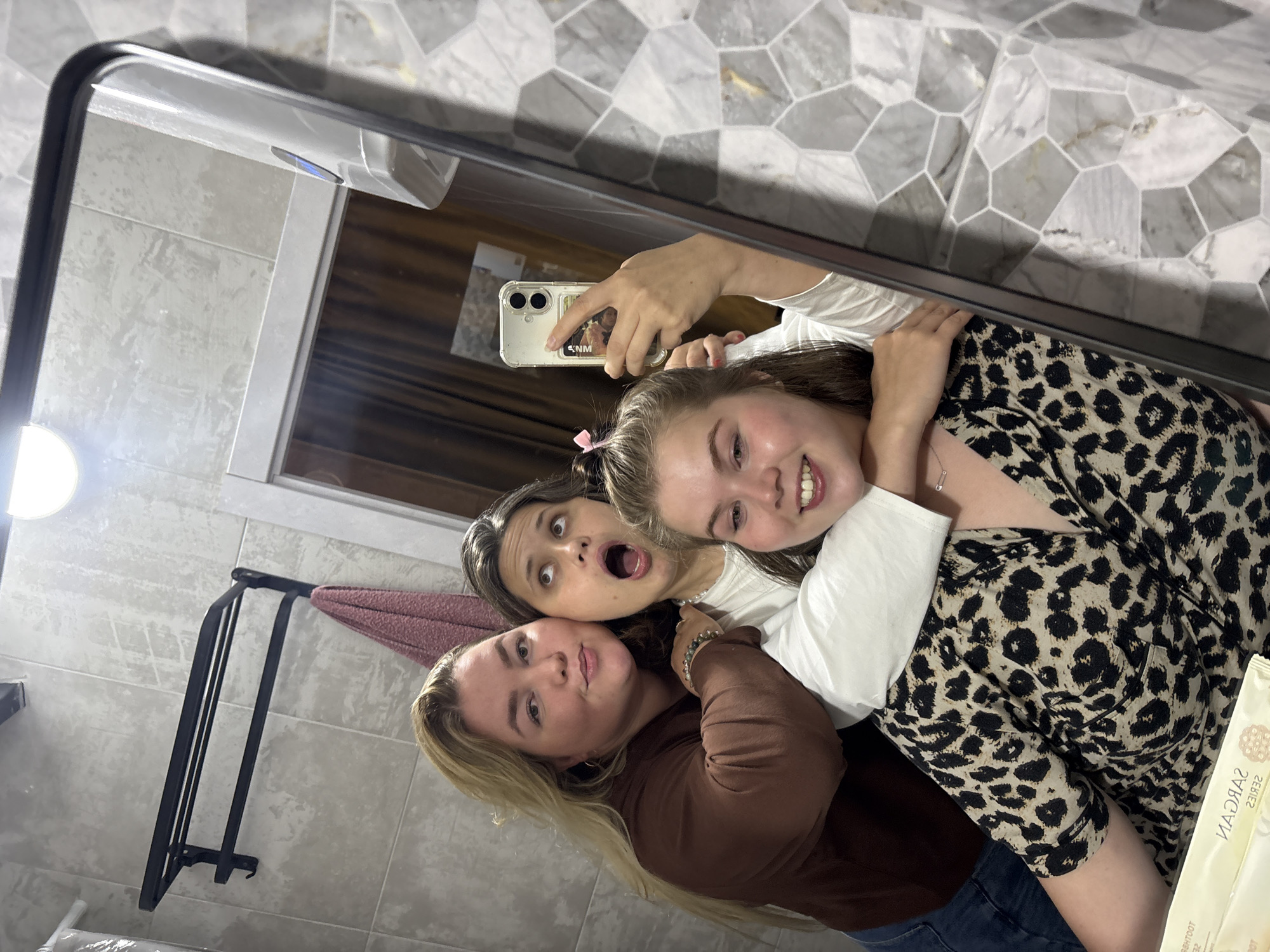 The image is a selfie taken in a bathroom. Three young women are posing for the photo. The woman on the left has blonde hair and is wearing a brown top. The woman in the middle has dark hair and is wearing a white top. The woman on the right has light brown hair and is wearing a leopard print top. She is holding a phone to take the picture. The bathroom has tiled walls and a mirror. A towel rack with a pink towel is visible on the wall. There is also a light fixture on the ceiling. The scene appears to be indoors, in a domestic setting. There is no information provided about the city of Костоваты, Russia.