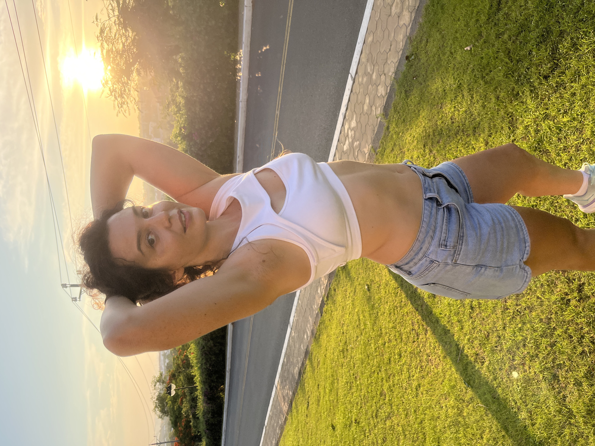 In the city of Phu Thuy Ward, Vietnam, a woman stands on a grassy area next to a paved road, with her arms raised and hands behind her head. She is wearing a white crop top with a cutout in the chest and light blue denim shorts. The sun is setting in the background, casting a warm glow over the scene. Trees and foliage are visible, along with utility poles and wires. The pavement beside the road has a patterned tile border.