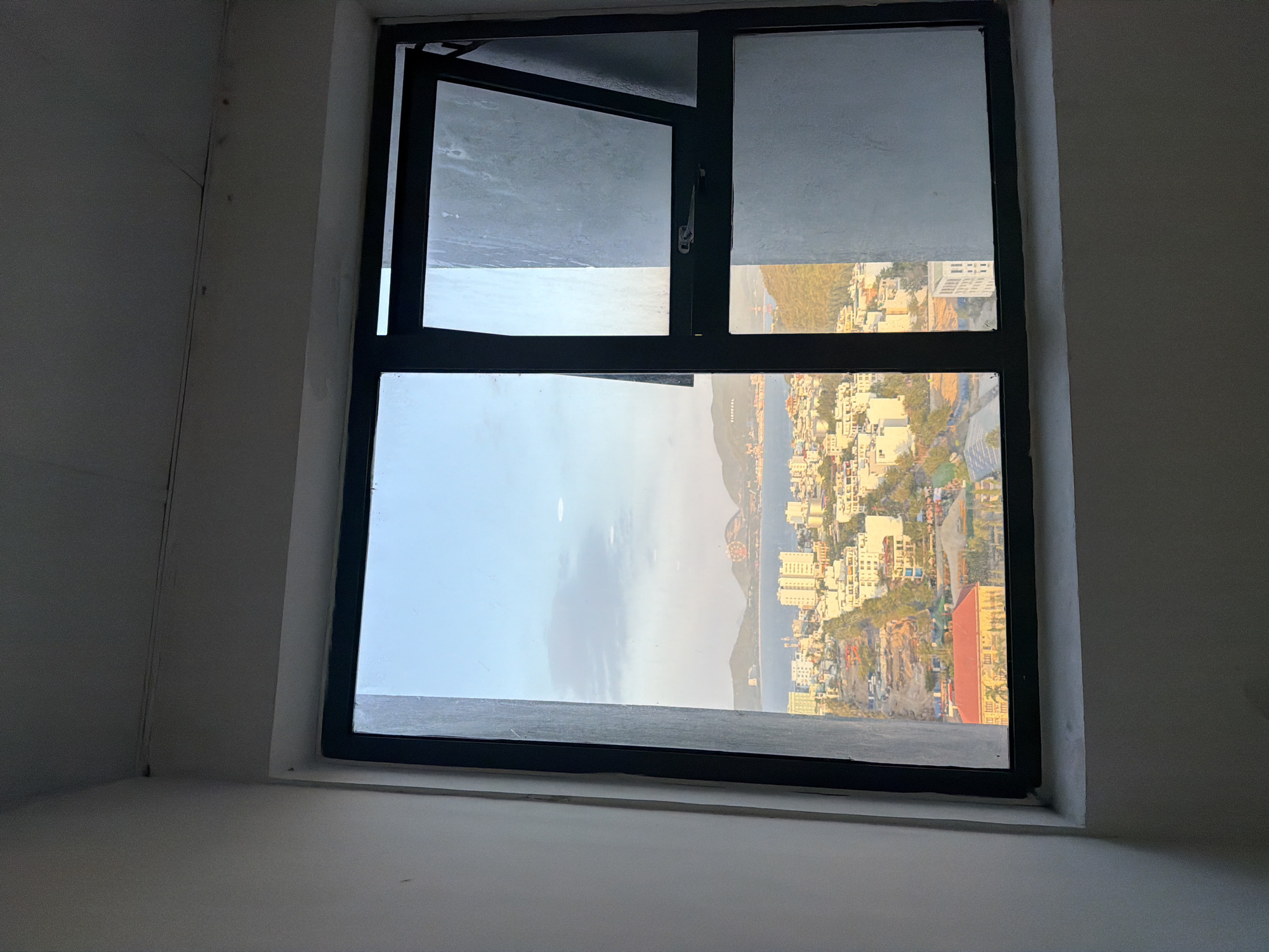 The image shows a view from a window overlooking the city of Nha Trang, Vietnam. The window is a double-paned casement window, with the right pane opened outwards at an angle. The interior of the room is dimly lit, with white walls. The view outside is a cityscape with numerous buildings of various heights, many appearing to be apartment buildings or hotels. A body of water, likely the sea, is visible in the distance, with hills or mountains on the far side. The sky is a clear blue with some scattered clouds, and the light suggests it is either early morning or late afternoon. There is a bridge visible in the distance over the water. There are no people or animals visible in the image. The scene appears to be a typical urban view from a high-rise building in a coastal city. The specific location within Nha Trang is not definitively identifiable without further context, but the presence of the sea, city buildings, and hills is characteristic of many areas within the city.