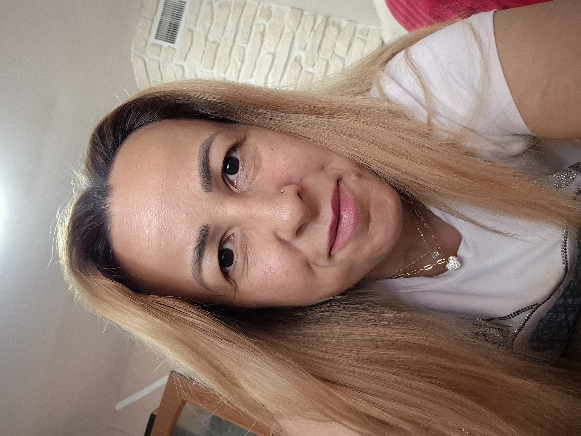 A medium shot shows a woman with blonde hair and fair skin looking directly at the camera. She has a faint smile and is wearing a white t-shirt with a jeweled neckline and two gold necklaces, one with a heart-shaped pendant. A nose ring is visible on her right nostril. The background is blurred and appears to be an indoor setting with a stone wall and ventilation grate on the right.