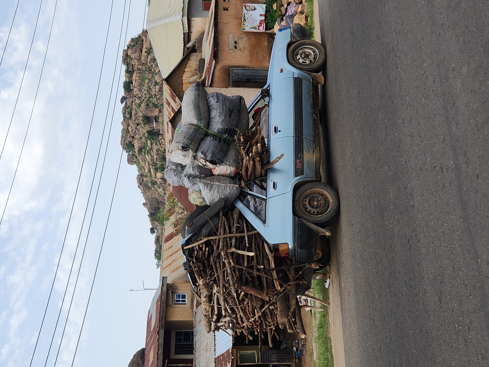 In Rukuba Road, Nigeria, a light blue car, heavily overloaded, sits on a paved road adjacent to a building. The car is laden with bags of what appears to be charcoal on top and a large quantity of firewood on the front. The tires of the car are worn. The building next to the car appears to be of simple construction with a rusty, corrugated metal roof. In the background is a rocky hill. Overhead are electrical wires spanning the sky. The road is asphalt and extends along the side of the image.