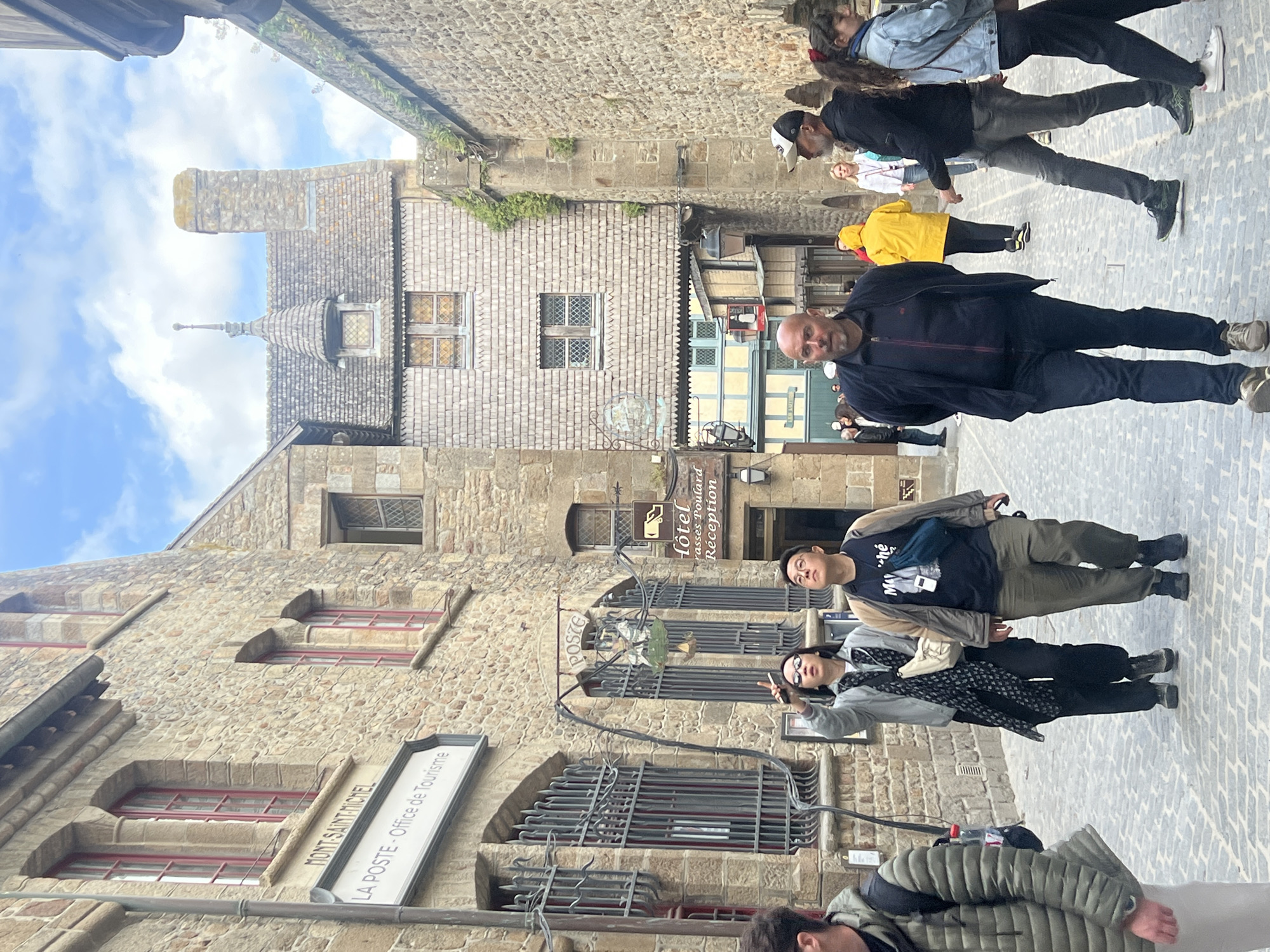 This detailed image captures a bustling street scene within Mont Saint-Michel, France, specifically near the Mont-Saint-Michel Post Office and Tourist Office, identifiable by the sign on the left-hand building.
The scene is set on a cobbled pathway flanked by ancient stone buildings under a partly cloudy blue sky. On the left, a multi-story stone building features arched windows with prominent red frames and metal grates on the lower windows. A white rectangular sign reads "MONT-SAINT-MICHEL" above "LA POSTE - Office de Tourisme." Below this, an ornate metal sign, partially obscured, appears to spell "POSTE" and displays an illustrative image. Further into the background, a distinctive building with a wooden shingle facade and multiple leaded-pane windows is visible, topped by a gray shingle roof featuring a small, pointed turret with a finial and a stone chimney. An archway passes beneath this building, revealing another structure with light blue and yellow sections. A sign for "Hôtel Mère Poulard Réception" hangs on the right side of the main wooden-shingled building. The right side of the street is bordered by a tall stone wall with patches of green foliage.
Several individuals populate the scene. In the foreground, the left side shows the torso and arm of a person wearing a green quilted jacket and a backpack. Slightly behind and to the left, a woman wearing glasses, a gray jacket, black pants, and a patterned black scarf holds a smartphone pointed upwards, seemingly taking a photograph or video. She has a light-colored crossbody bag. Next to her, a man with glasses, a dark t-shirt, a light brown jacket, and dark pants looks upwards. He carries a blue fanny pack and a white rectangular device on his chest. Walking towards the viewer in the center is a bald man with a short beard, wearing a dark blue zip-up jacket over a dark t-shirt and dark blue jeans. To the right, a person in a bright yellow hooded raincoat walks away from the viewer. To their right, a man wearing a white and dark baseball cap, a dark jacket, and grey jeans walks with his head turned slightly to the right. Beside him, a woman with long dark hair, wearing a light blue denim jacket and dark pants, walks away from the viewer. Additional pedestrians are visible further down the street in the background.