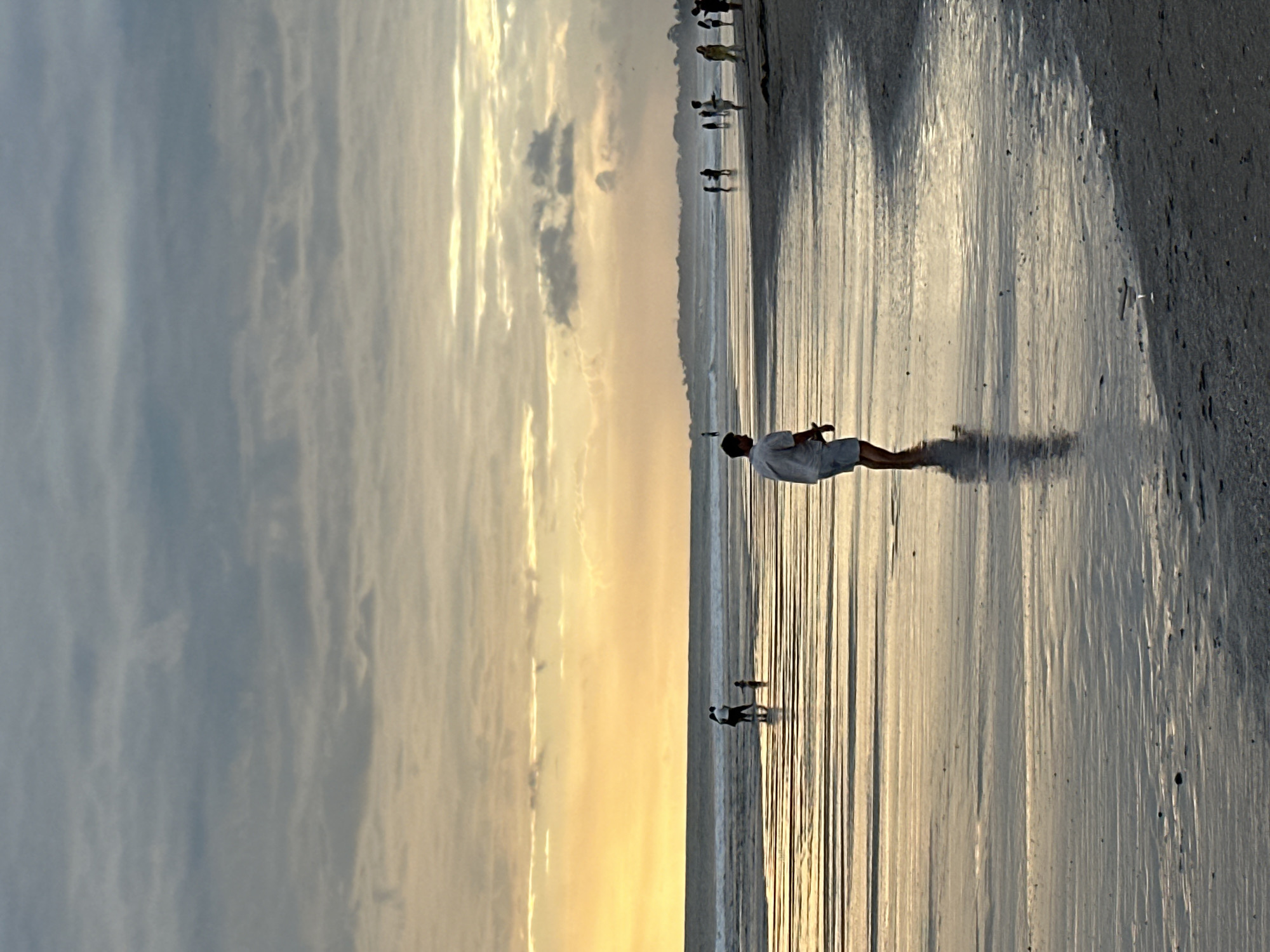 A wide shot captures a serene sunset scene at the beach in North Kuta, Indonesia. The sky is filled with a blend of muted gray and soft orange hues, indicating the sun is setting. The ocean is calm with gentle waves rolling towards the shore. The wet sand reflects the colors of the sky, creating a glistening effect with horizontal lines of light. Several figures are scattered across the beach. In the foreground, a lone person in a white shirt and blue shorts stands facing away from the camera, their reflection visible in the shallow water. Further down the beach, a group of people are walking near the water's edge, and another small group is further out in the sea. The overall atmosphere is tranquil and peaceful, with the natural beauty of the sunset dominating the scene.