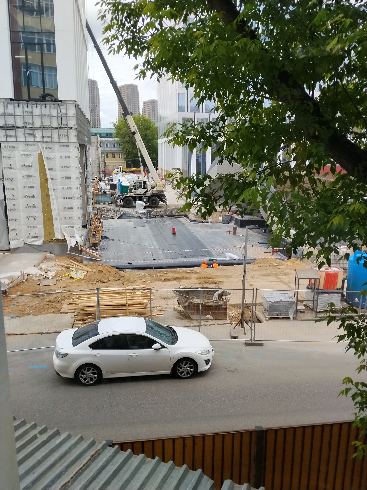 Here's a detailed description of the image:
**Scene Overview:**
The image captures a view of a construction site within an urban environment, likely in Moscow, Russia. The scene is observed from an elevated position, possibly from a building window or balcony.
**Foreground:**
* A white four-door sedan (possibly a Mazda) is parked on a paved road.
* Below the viewing position, corrugated metal and a wooden fence form part of the infrastructure.
* A fence separates the road from the construction zone.
**Construction Site:**
* Significant construction activity is taking place.
* Construction materials, including piles of wood, metal containers, and concrete blocks, are scattered around.
* Heavy equipment like a large mobile crane is present. The crane's boom extends upwards, suggesting ongoing operations.
* A dark-colored, likely waterproof, material is spread on a prepared section of the ground.
* Two people in orange vests are at the center of the construction site.
**Buildings:**
* Modern buildings dominate the background. These structures feature contemporary architectural styles with extensive use of glass and paneling.
* One building's facade appears to be partially covered with white material labeled "TEND" repeatedly.
**Background and Environment:**
* Skyscrapers are visible in the distance.
* The presence of trees with green foliage suggests a warm season.
* The sky is overcast.