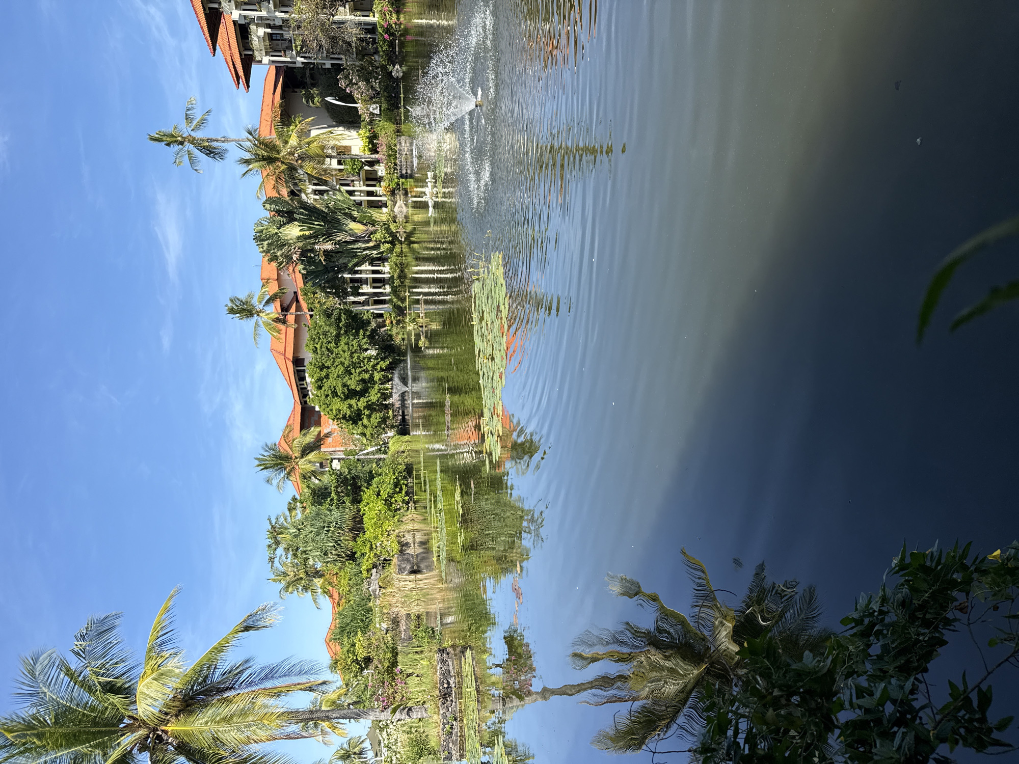 The image depicts a pond surrounded by lush greenery and buildings with red-tiled roofs, likely located in Kuta Selatan, Indonesia. A fountain is visible in the pond's center. The water reflects the sky, trees, and structures, creating a mirror-like effect. Palm trees are prominent features, adding to the tropical ambiance. No people are visible in the frame.