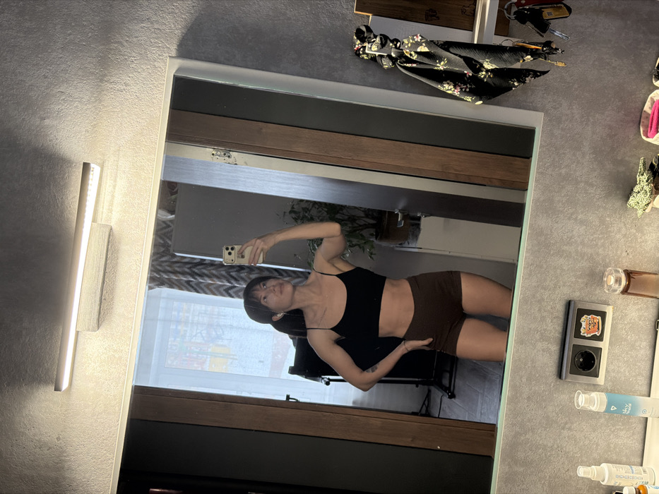 The image depicts an adult female capturing a mirror selfie in an indoor setting.
**Location:** The image provides no visual information to identify the location as Perm, Russia.
**Scene Description:**
The primary subject is a female, seen in a large rectangular mirror reflection. She has dark hair styled with bangs and pulled back into a ponytail, and is wearing wireless earbuds. Her attire consists of a black spaghetti-strap crop top and dark brown fitted shorts. A faint tattoo is visible on her left upper arm. She poses with her left hand on her hip, while her right arm is raised, holding a light-colored smartphone (identifiable by its camera array) towards the mirror to take the photograph.
The mirror is bordered by a white frame on the bottom and right, and a dark wooden frame on the left. Above the mirror, a horizontal LED light fixture is mounted, providing illumination. The reflection in the mirror shows elements of the room behind the subject, including a window with sheer white curtains, a patterned dark curtain, a large dark potted plant, and a black-framed stand or piece of equipment.
Below the mirror, a grey speckled surface, consistent with a countertop or wall, is visible. Arranged on this surface, from left to right, are: several bottles, including a small amber dropper bottle and a white bottle with partial text "Sky Natural"; a double electrical power outlet with two circular sockets, one adorned with a small cartoon pot-plant sticker; and a wooden key organizer or hook rack on the far right, from which a black floral-patterned scrunchie and multiple keys are hanging.