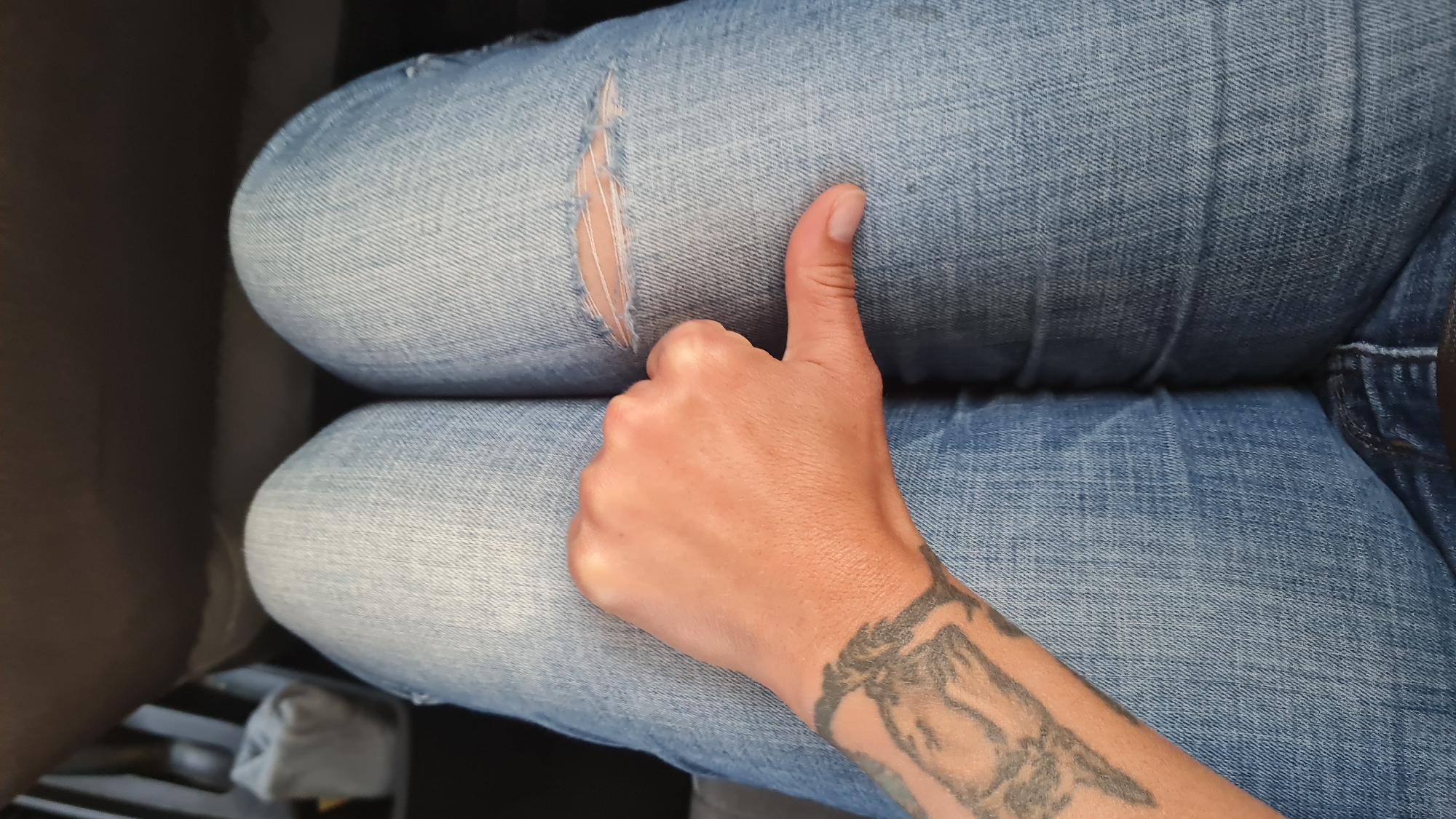 A person sits in what appears to be a car seat. They wear blue denim jeans with a rip at the upper thigh of their left leg. The person's arm, with a tattoo on the wrist, rests on their legs, giving a thumbs-up. The location is Bnei Shimon Regional Council, Israel.