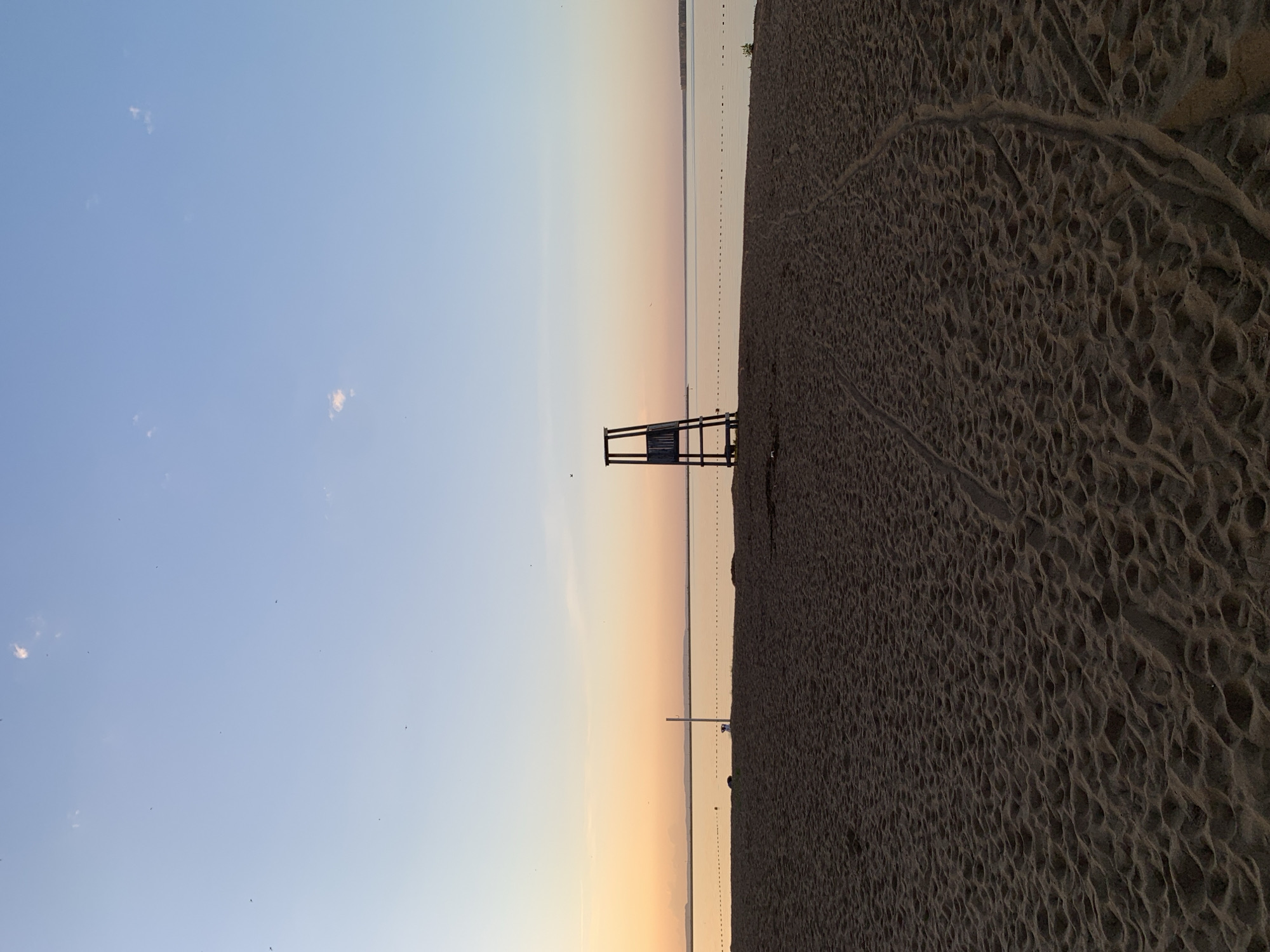The image depicts a beach scene at sunset in Chaikovsky, Russia. The sky transitions from blue to orange and pink hues. A lifeguard tower stands on the sand, silhouetted against the sunset. A body of water stretches out towards the horizon, with a line of buoys visible. The beach sand shows patterns of footprints and natural textures.