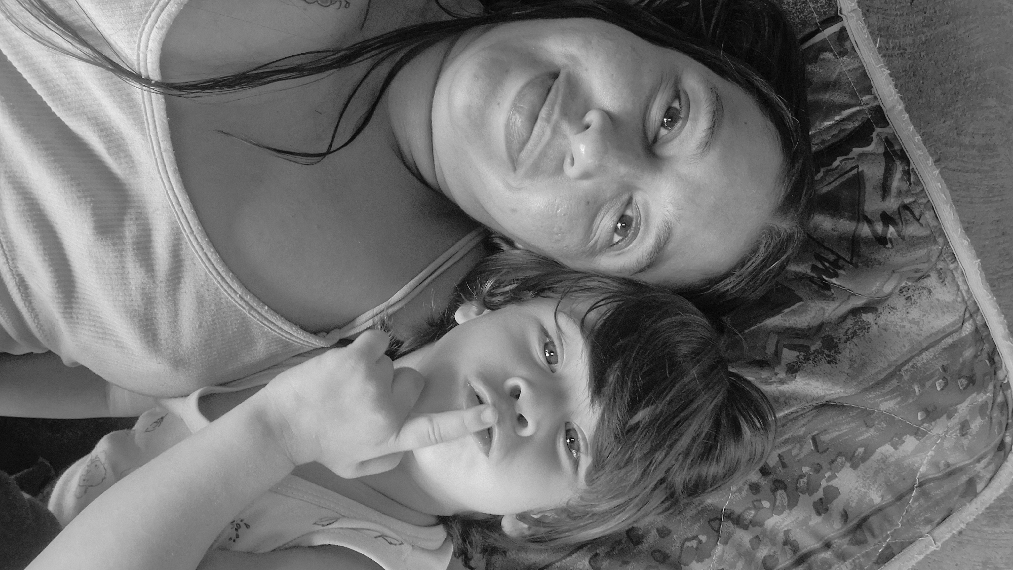 A black and white, close-up, overhead shot shows a woman and a young child lying down together. The woman is on the left, looking at the camera with a slight smile. She has long, dark hair and is wearing a ribbed tank top. The child is on the right, with their finger to their lips as if telling a secret. The child has short, dark hair and is wearing a light-colored tank top with a subtle pattern. They are lying on a patterned surface, possibly a bed or blanket. The overall lighting is soft, creating a natural feel. The image appears to be taken indoors, in what could be a bedroom or living space. The location of Vallecito, Venezuela is not discernible from the visual information in the image.