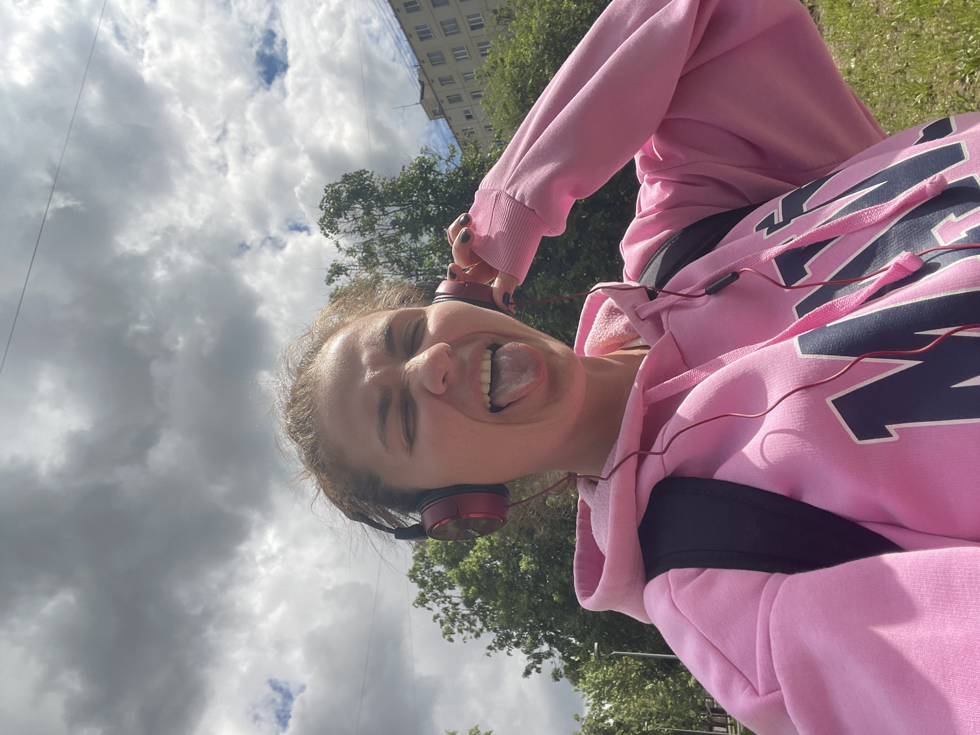 A young woman in a pink hoodie and wearing headphones is photographed from a low angle. She is sticking out her tongue and has her eyes squinted. In the background, there are trees, a building, and a cloudy sky. This scene is likely in Moscow, Russia based on the architecture of the building in the background.