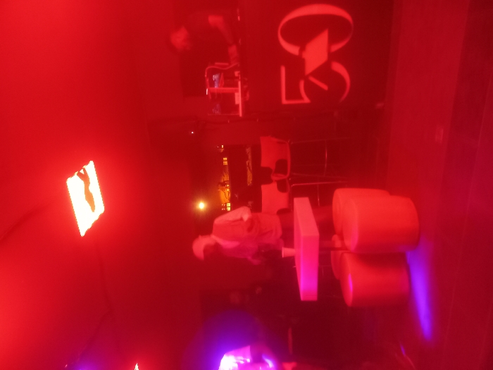 A red-lit interior features a seated person, likely in a spa or treatment setting. The room has a chair with cylindrical cushions and a nearby light fixture on the wall. A logo is seen on the wall, with a person in the background tending to equipment. The overall ambiance is dark and appears to be in a wellness or relaxation space.