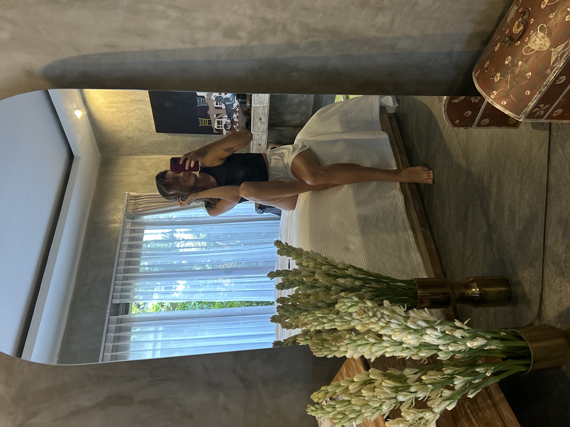 This media file is a mirror selfie taken by a woman in what appears to be a modern bedroom or hotel room. The image is oriented vertically but depicts a horizontal scene.
The central subject is a woman, reflected in a large, slightly rounded mirror. She has dark hair, is wearing a black tank top and white shorts, and is lying or reclining on a bed. One leg is bent at the knee, while the other is extended. She holds a red smartphone with both hands, pointing it towards the mirror to capture her reflection. Her face is partially obscured by the phone.
The room features concrete-like grey walls and a white ceiling with recessed lighting. The bed has a light-colored wooden frame and is covered with white, textured bedding. Behind the bed, a dark rectangular piece of artwork hangs on the wall, depicting light-colored figures that resemble buildings or cats. A wooden bedside table or dresser is also visible. To the right of the bed, a patterned, upholstered chair or piece of furniture is partially seen.
In the foreground, two bouquets of long, white and pale green flowers (possibly tuberoses) are arranged in brass-colored vases, placed on a wooden surface. A large window with sheer white curtains allows natural light into the room, revealing glimpses of lush greenery outside. The flooring appears to be a dark, textured material.
The overall impression is one of a contemporary and relaxed setting, likely in North Kuta, Indonesia, as suggested by the context. There is no discernible text visible in the image. The time of day appears to be daytime due to the light entering through the window.