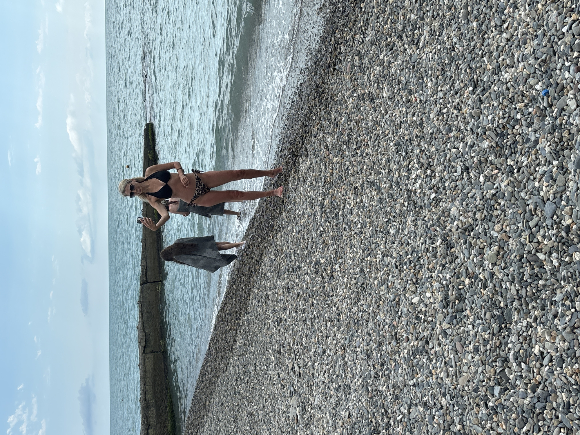 The image shows a beach in Sochi, Russia, with a woman in a black bikini and leopard print bottoms taking a selfie with her phone. She is standing in shallow water on a pebble beach, with the sea in the background. Another person, wearing a grey cover-up, is standing to her left, and further to the left, another person in the water. A stone breakwater extends into the sea. The sky is partly cloudy.