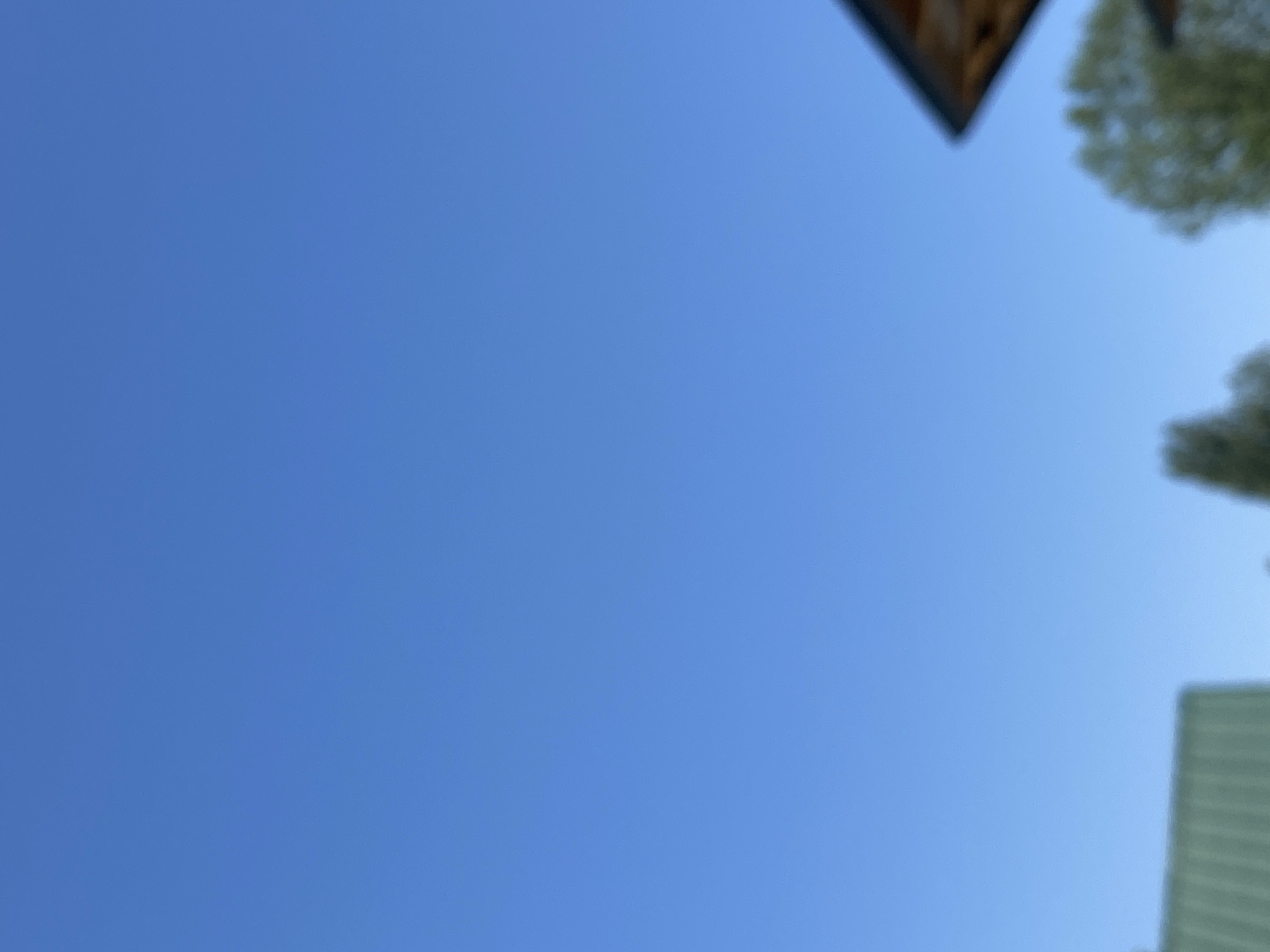 The image shows a clear blue sky. In the bottom left corner, a portion of a green roof of a building is visible. To the right of that, out-of-focus trees are present. In the upper right corner, the edge of a wooden roof or structure is visible. The scene appears to be outdoors, with no specific identifiable landmarks to pinpoint it to Arbagish, Uzbekistan.