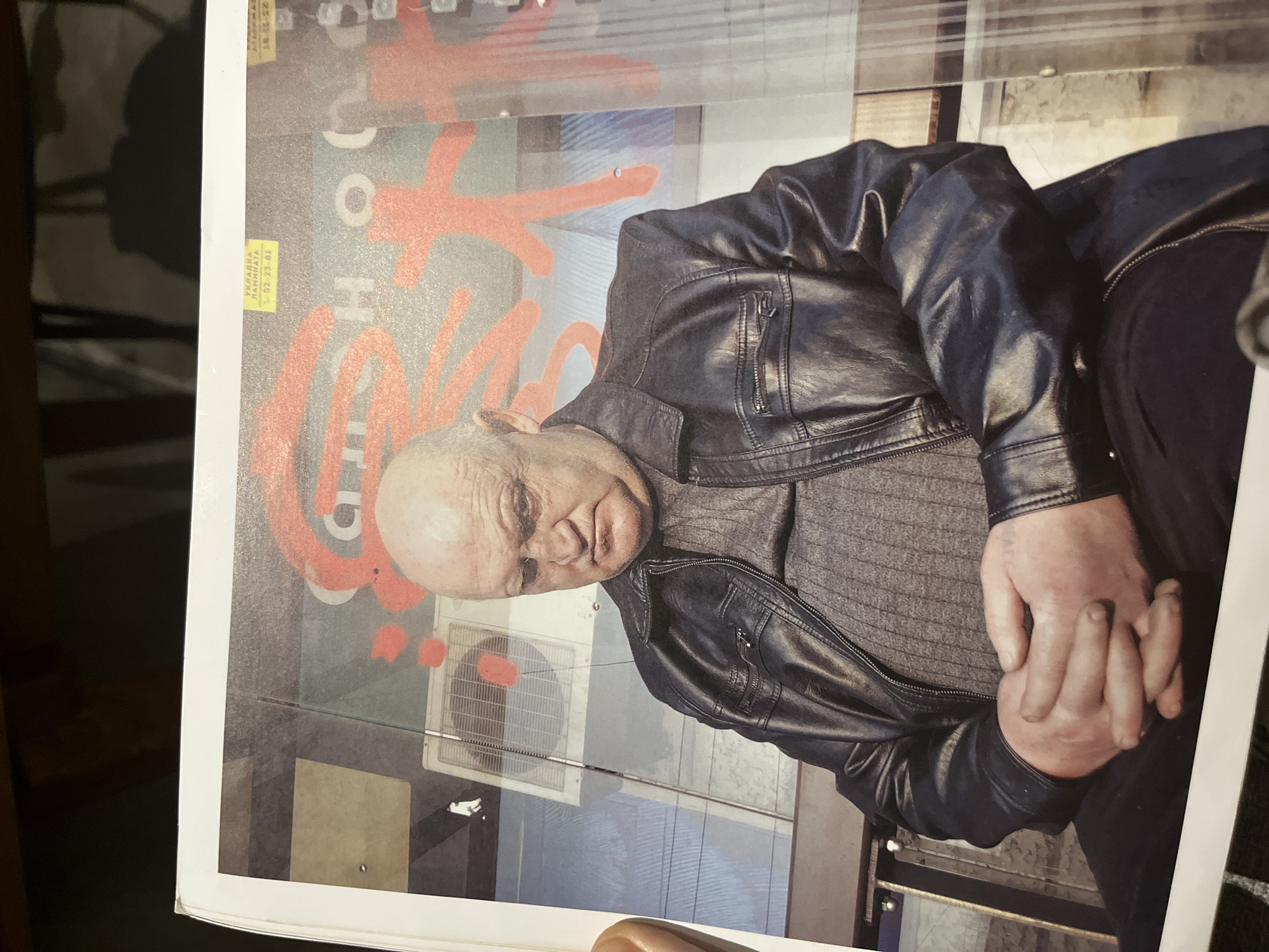 A photograph, printed on paper, displays a bald man wearing a dark leather jacket over a gray sweater. He is sitting, with his hands clasped in front of him. Behind him is a building facade with graffiti, an air conditioning unit, and signage. The text on the signage appears to be in Cyrillic script, with "УКЛАДКА ЛАМИНАТА" and a phone number visible. The image is rotated counter-clockwise.
I am unable to provide the specific location in the Ubud District, Indonesia without additional information.
