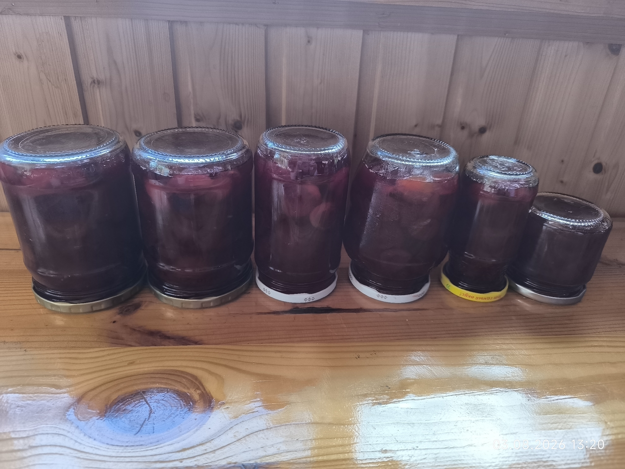 A slightly high-angle, medium shot shows six glass jars filled with a dark red preserved food, likely jam or compote, arranged in a line on a wooden surface. The jars vary in size and shape, with some taller and narrower than others. They are capped, with some lids appearing to be metal and others possibly metal with a white rubber seal. The wooden surface appears to be a planked table or shelf, with visible grain and a glossy finish. Behind the jars is a wooden plank wall, also with visible grain. The lighting is moderate, casting some subtle shadows. A timestamp in the bottom right corner reads "03.08.2026 13:20", suggesting a date in the future. The location is identified as "Чэрвеньскі сельскі Савет, Belarus". There are no people or animals visible in the image.