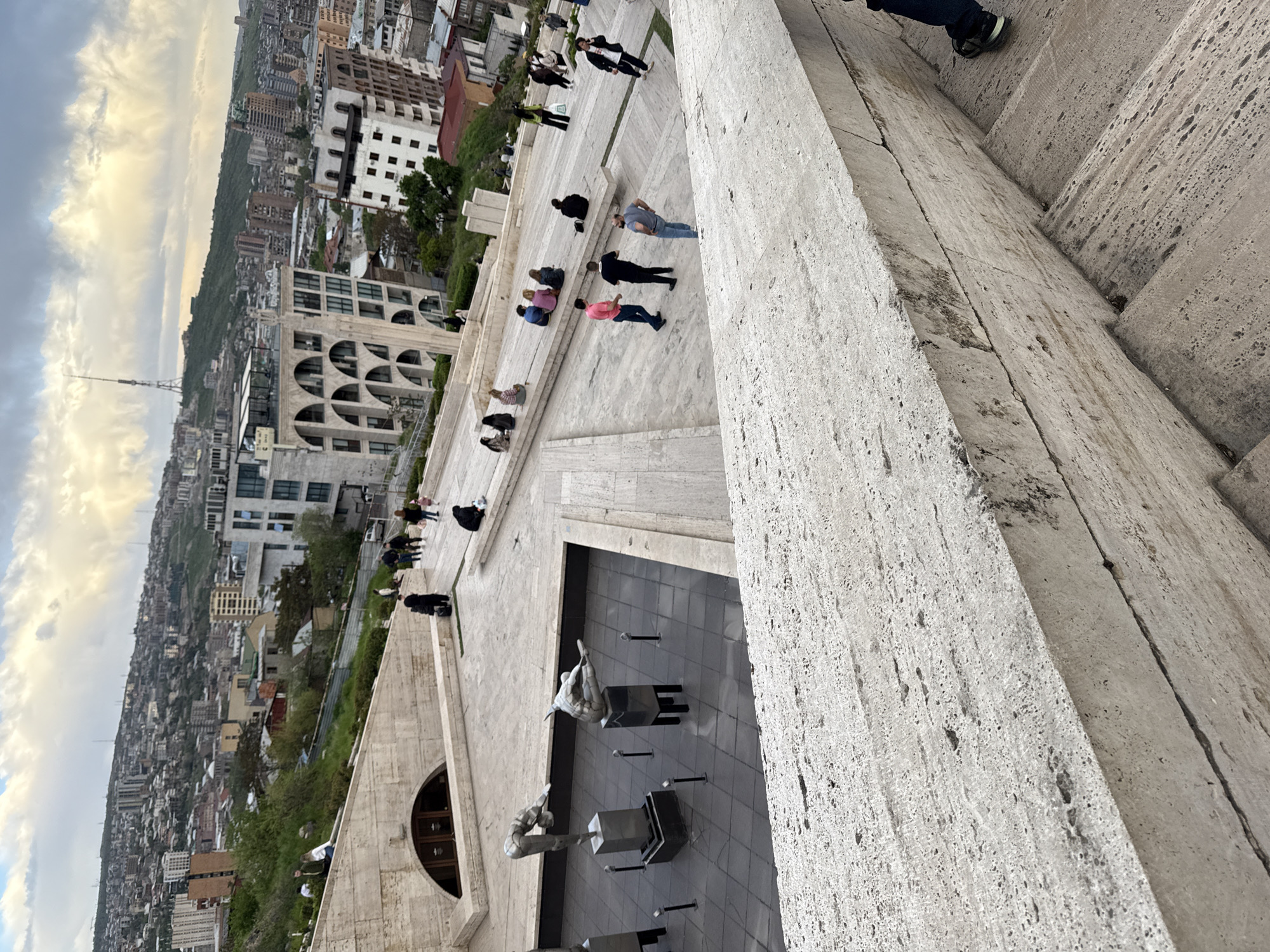 This image captures an elevated view of the Yerevan Cascade in Yerevan, Armenia.
People are spread across the steps of the Cascade, some standing, others sitting and resting. The cityscape of Yerevan extends into the background under a sky with clouds. Sculptures are visible at one of the lower levels. The architecture of the Cascade is prominently displayed, with its light-colored stone steps.