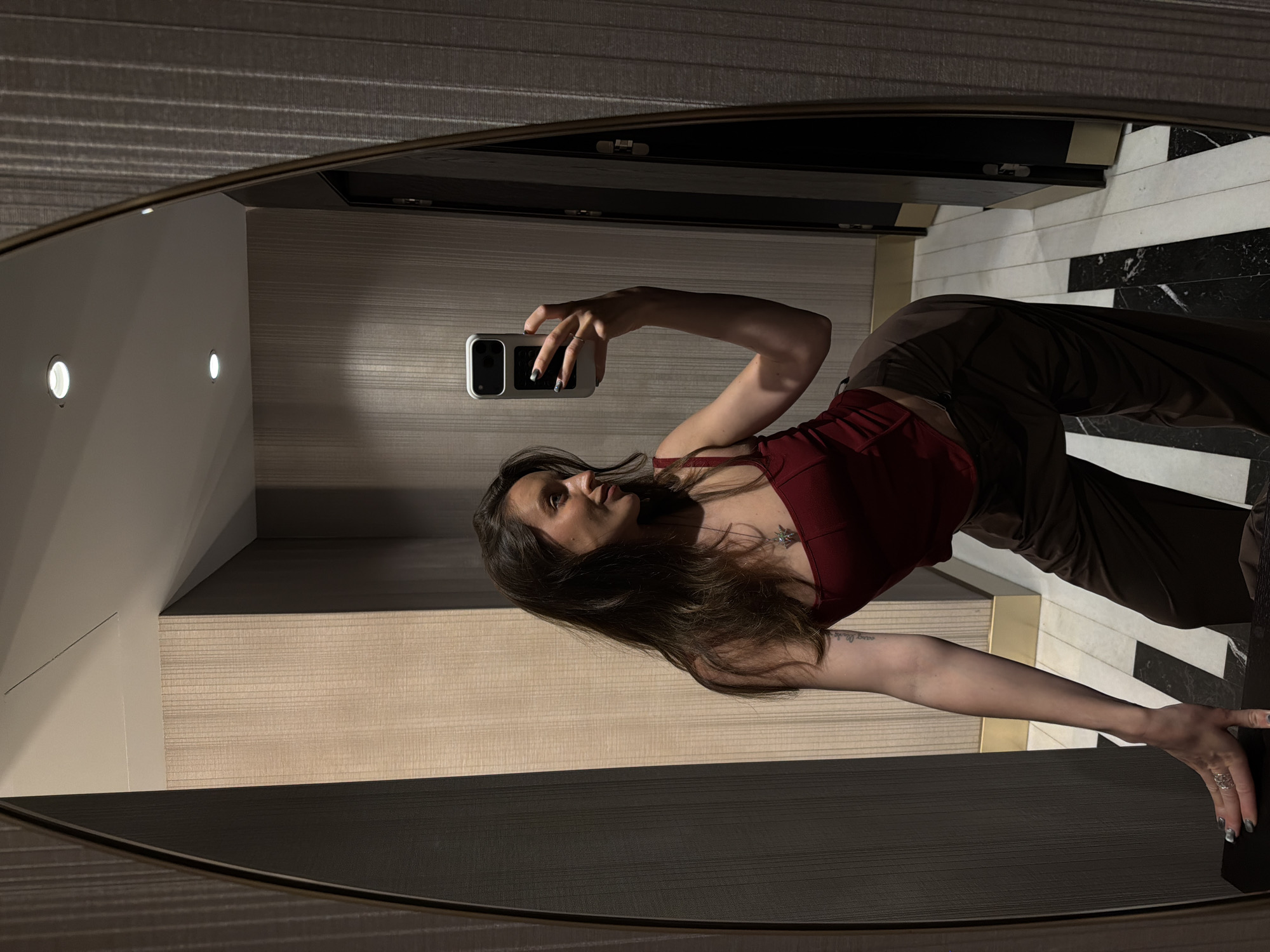A woman is taking a selfie in a mirror. The reflection in the mirror shows a portion of a hotel room. The walls are beige. The floor is black and white marble. The woman is wearing a red top and dark pants. It is not possible to determine the location of the hotel room specifically in Budva, Montenegro from this picture.
