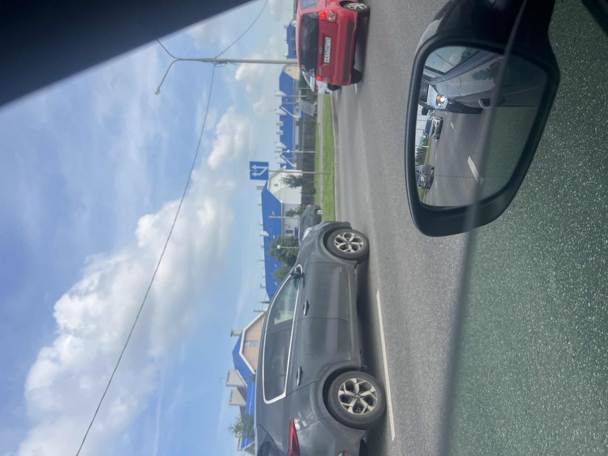 The image depicts a street scene in Magnitogorsk, Russia, on a cloudy day. In the foreground, a dark grey SUV is visible, positioned on a road with a white line marking the lane. To the right, a side-view mirror reflects several cars in traffic, including a red hatchback with a license plate that appears to read "K417HB 174". In the background, a row of modern houses with distinctive bright blue roofs is lined up along a street. A blue and white traffic sign indicating lane directions is also visible. The sky is partly cloudy, with scattered white clouds against a light blue backdrop. The overall environment suggests a suburban or newly developed residential area within the city.