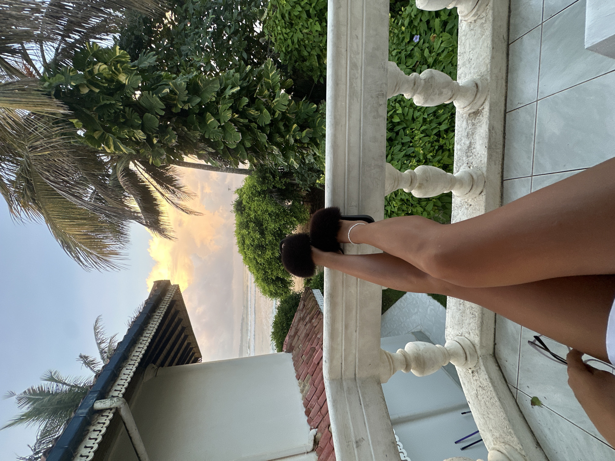 The image depicts a person's legs and feet resting on a white balcony railing, with the ocean and lush greenery visible in the background. The person is wearing fuzzy dark slippers and a delicate anklet. The balcony features decorative balusters and a tiled floor. The scene suggests a tropical or subtropical setting, likely a vacation spot, possibly in Peleana, Sri Lanka, given the presence of palm trees and the coastal view. The sky shows soft, warm clouds, indicating either sunrise or sunset.