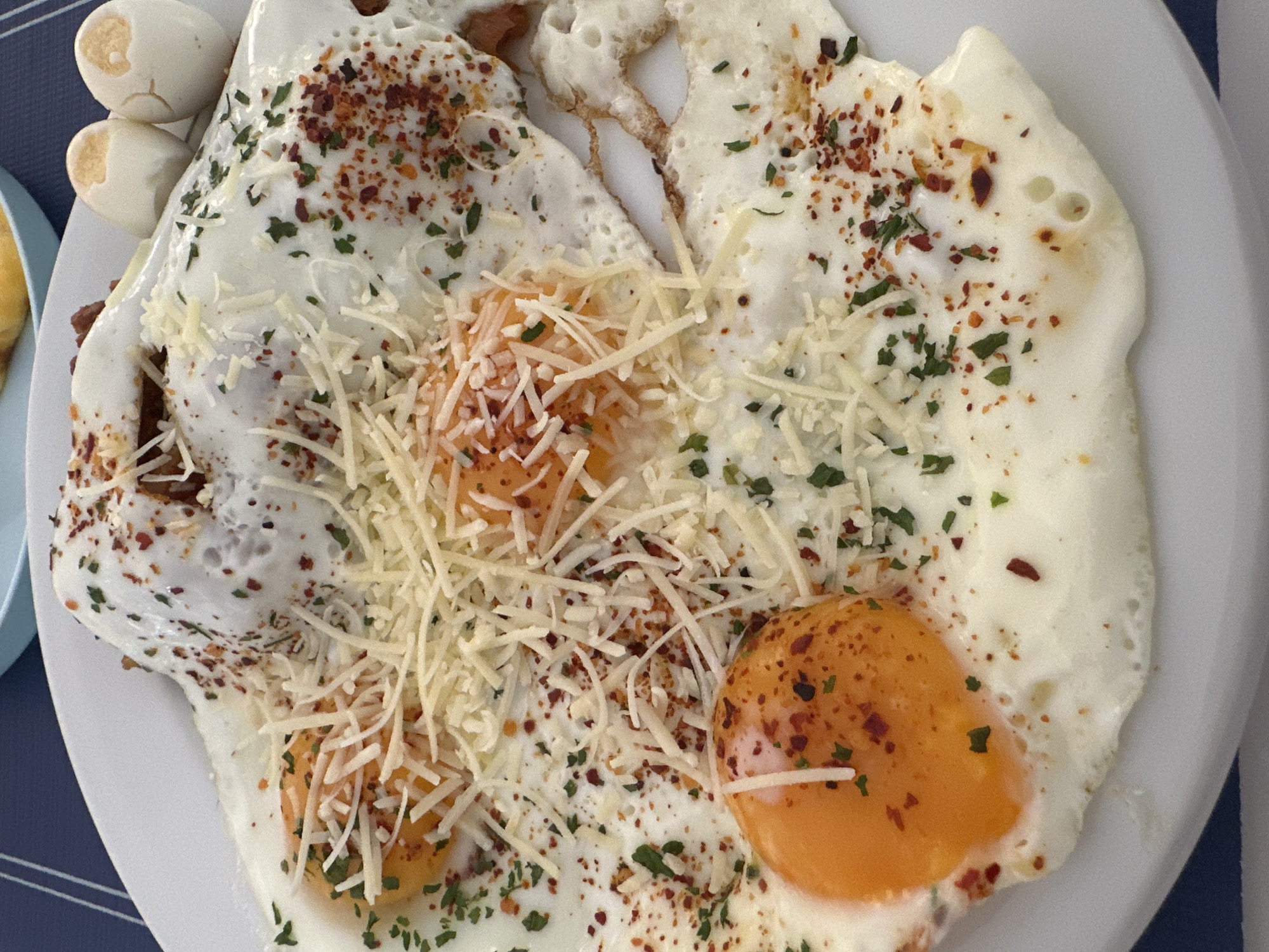A close-up, overhead shot shows a white plate filled with fried eggs, some with runny yolks, sprinkled with shredded cheese, dried herbs, and red pepper flakes. To the upper right, two hard-boiled eggs with cracked shells are visible. The plate rests on a blue and white striped tablecloth. The scene is likely from a breakfast setting in the city of Sant Feliu de Guíxols, Spain.