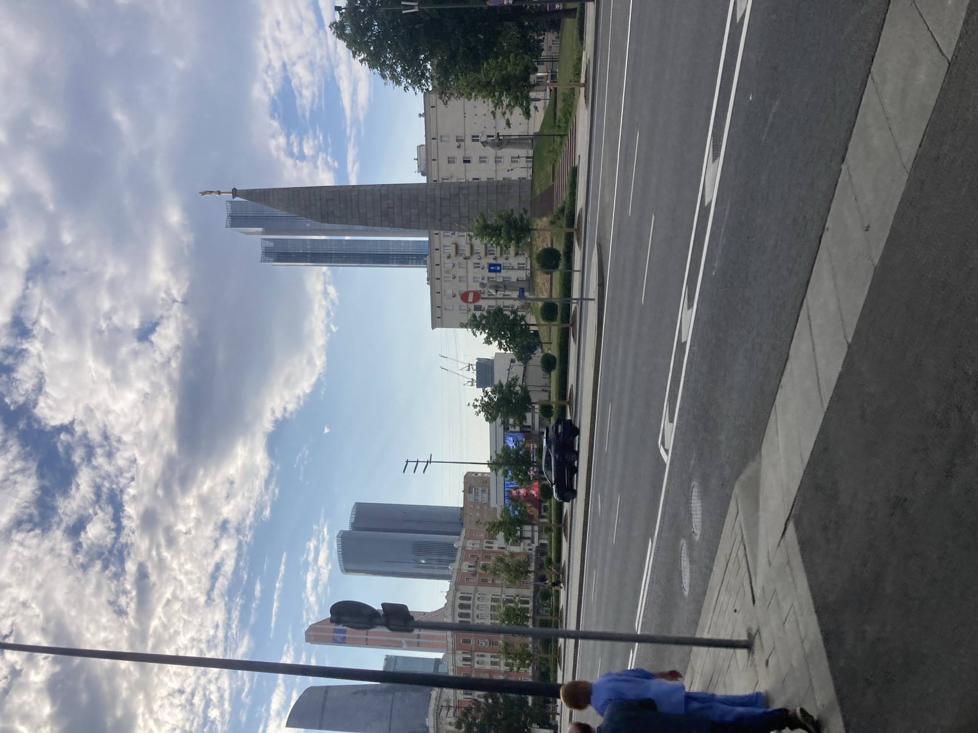 This image captures a street scene in Moscow, Russia, under a partly cloudy sky, suggesting it's daytime with fair weather. The perspective is from street level, looking towards a row of modern and older buildings, a road with traffic, and a sidewalk.
Prominently featured are several tall buildings, including two sleek, modern glass skyscrapers and a larger, imposing concrete structure that appears to be a monument or an older, monumental building. In the mid-ground, there's a building with a more traditional architectural style, adorned with arched windows. A dark-colored car is visible on the road.
On the sidewalk in the foreground, a person dressed in blue clothing is seen with their back to the camera, appearing to be walking or standing. The overall atmosphere is that of an urban environment with a mix of contemporary and historical architecture. There is no visible text in the image.