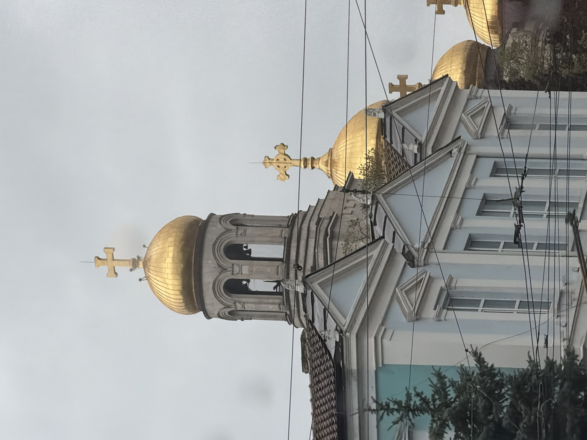 This image displays an upward-looking view of an Eastern Orthodox church, fitting the architectural style found in Varna, Bulgaria. The dominant features are the numerous gleaming golden domes, each crowned with a distinctive Orthodox cross. The largest dome sits atop a bell tower section, characterized by arched openings. The church's facade is light-colored, possibly pale blue or white, adorned with decorative elements including triangular pediments and ornate moldings around windows and structural lines.
The sky is uniformly overcast and light gray, suggesting a cloudy or dull daytime. Numerous dark electrical wires crisscross prominently across the frame, obscuring parts of the sky and the church itself. Dark green tree foliage is visible in the lower and right portions of the image (when correctly oriented), partially obscuring the lower sections of the building. There are no people visible, nor is there any discernible text. The image captures a blend of traditional religious architecture with the presence of modern urban infrastructure.