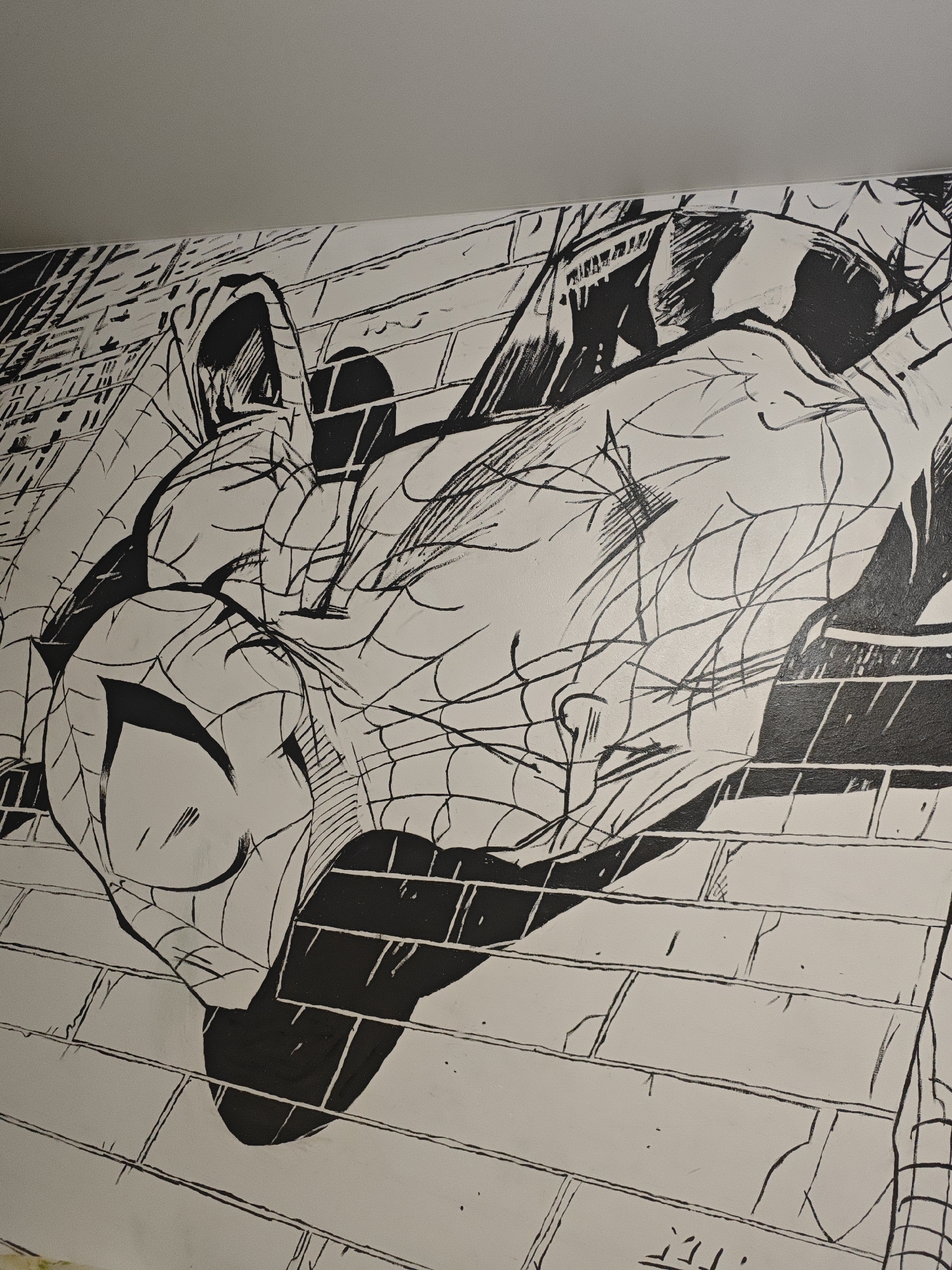 The image shows a black and white mural on a brick wall. The mural depicts Spider-Man in a dynamic pose, with webs drawn across his body. The background of the mural appears to be a city skyline with buildings. The mural is done in a comic book style with bold lines and strong contrasts between black and white. The location of this mural is unknown, and it is not possible to determine if it is located in O'lkhovka, Russia based on the image alone.