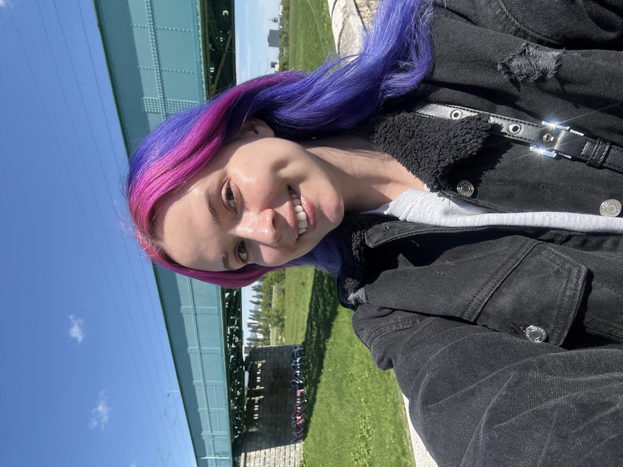 A young woman with vibrant, multicolored hair smiles at the camera. Her hair is predominantly purple and pink, cascading across her head and shoulders. She is wearing a black denim jacket with a furry collar and a white t-shirt underneath. A silver studded belt is visible on her jacket.
The background shows a sunny outdoor scene, likely a park area. A green metal bridge spans the landscape, and a grassy area stretches behind the subject. In the distance, there are trees and buildings, indicating an urban setting. Graffiti is visible on a stone structure beneath the bridge.
Given the presence of the bridge and the grassy terrain, this image likely depicts the underpass of the Hendrix bridge near Bundek Lake in City of Zagreb, Croatia.