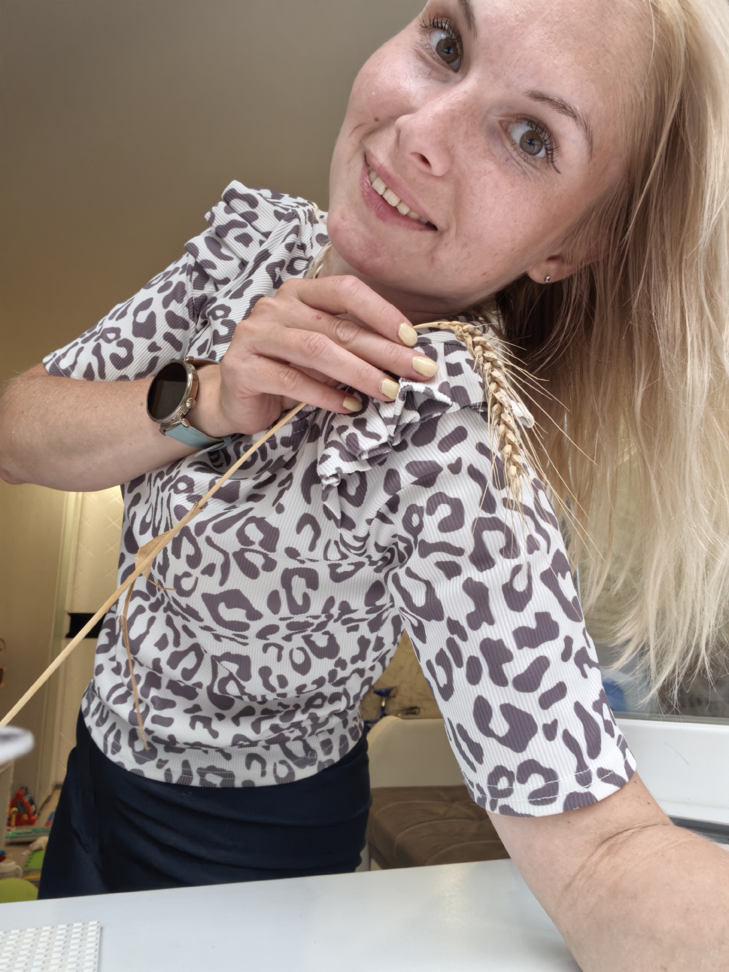 A woman with blonde hair and fair skin is looking at the camera with a smile, holding a stalk of wheat. She is wearing a white shirt with a grey leopard print and a black skirt. A smartwatch is visible on her left wrist. The background is blurred, but appears to be an indoor setting with some natural light. There are no identifiable landmarks or context to suggest the location in Sterlitamak, Russia.