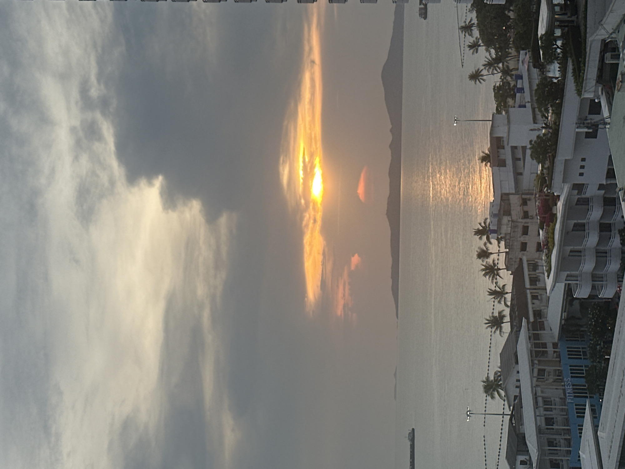 View of Pattaya City, Thailand. Cloudy sky with a break in the clouds revealing a sunset. Buildings on the right edge of the image, a beach, palm trees, and a body of water. The sun's reflection is visible on the water. A hill is on the horizon.